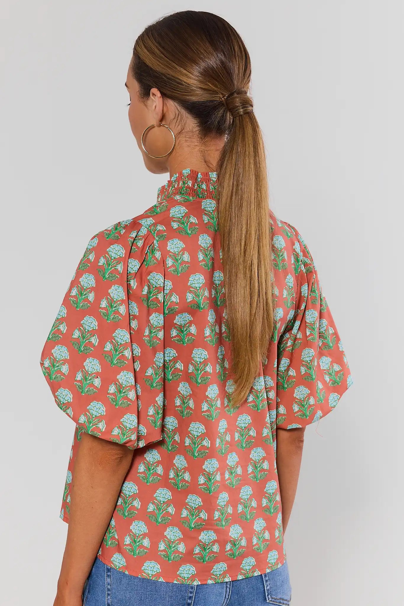 A woman wearing a coral-orange short-sleeve blouse with a green and light blue floral block print and a cream-colored split neckline shown from the back