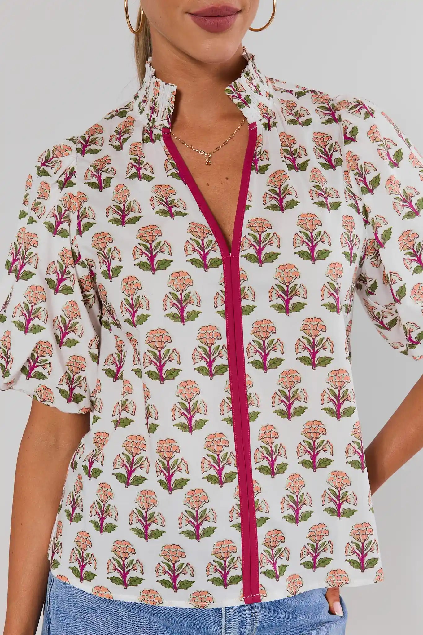 Close up of a woman wearing a short-sleeve button-up blouse with a white base, pink and green floral block print, and a contrasting bright pink front zipper, paired with blue jeans against a light background.
