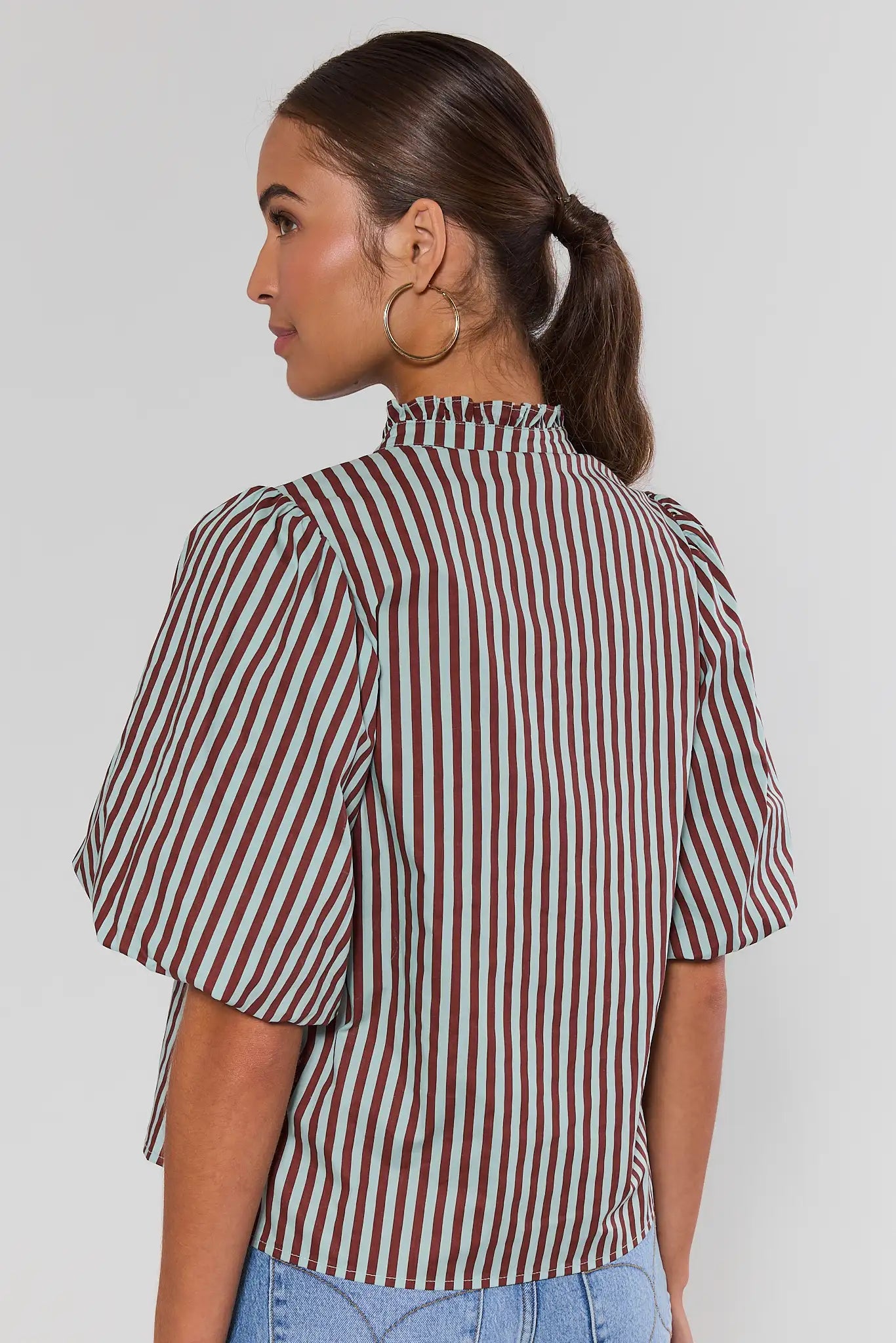 A woman shown from behind wearing a short-sleeve button-front blouse with vertical burgundy and mint green stripes, paired with gold hoop earrings, against a plain gray studio background.
