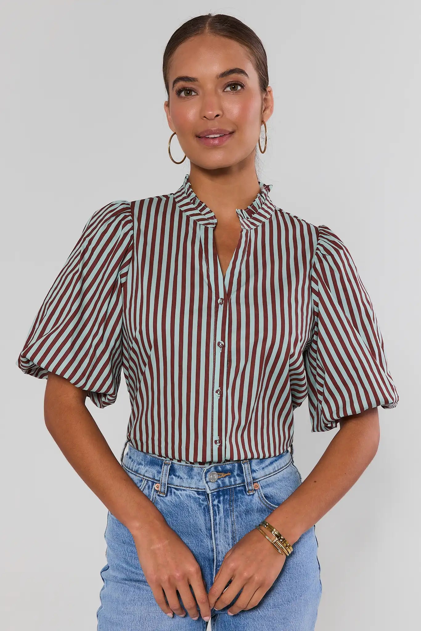 A woman wearing a short-sleeve button-up blouse with vertical stripes, paired with light blue jeans, against a plain light gray background.