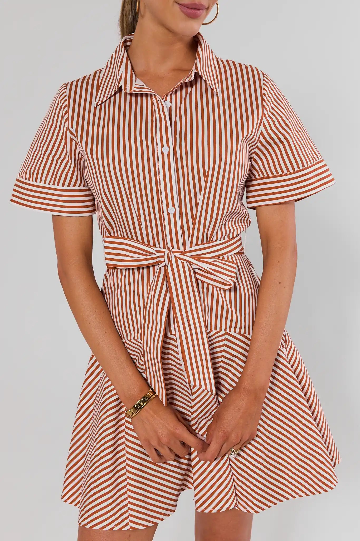 A woman wearing a short-sleeve collared shirt dress with vertical rust-orange and white stripes, featuring a button front, tie waist, and flared skirt close up