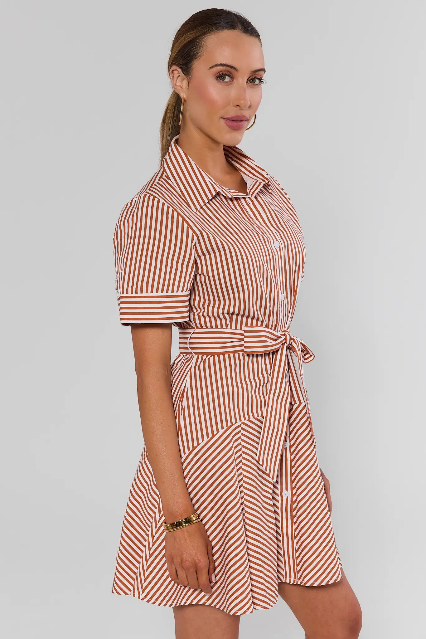 A woman modeling a short-sleeve collared shirt dress with vertical red and white stripes, a button-up front, and a matching tie belt at the waist, photographed against a light studio background 