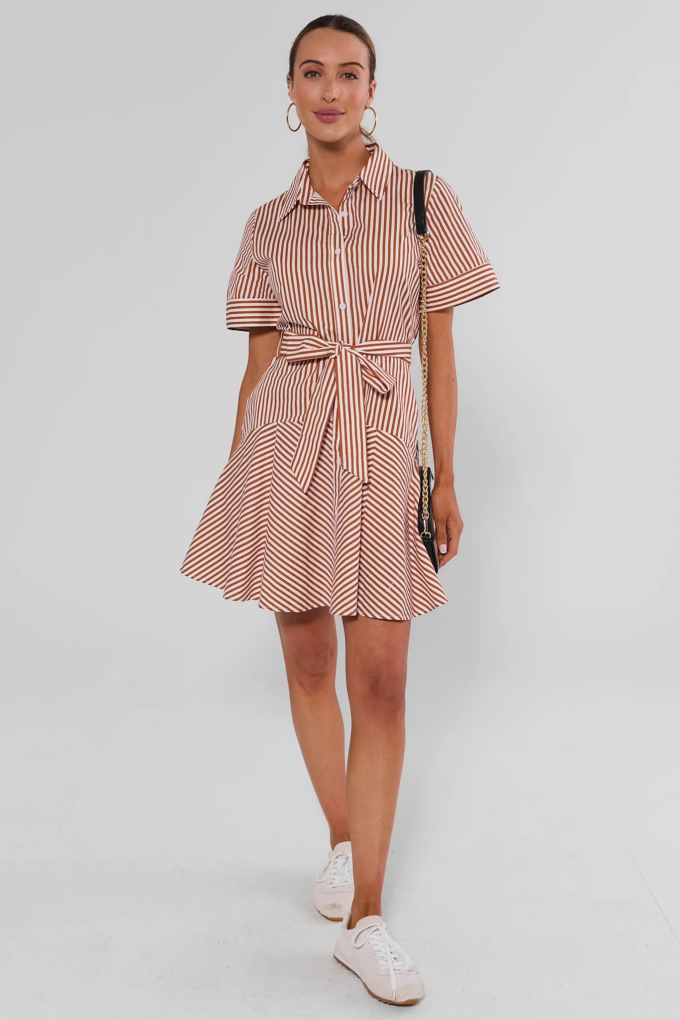 A woman modeling a short-sleeve collared shirt dress with a vertical pink and white stripe pattern, featuring a tie waist and flared skirt, paired with white sneakers and a black shoulder bag against a light gray studio background.