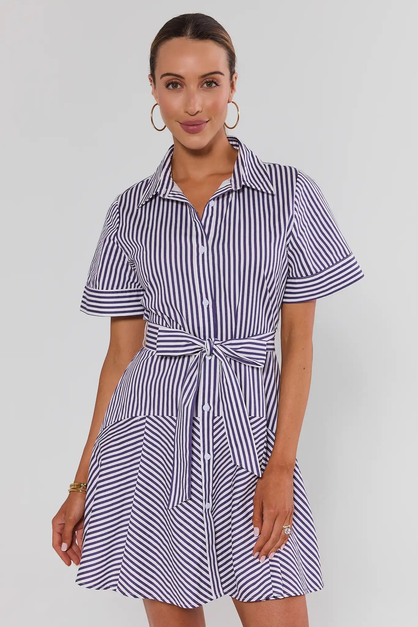 A woman standing wearing a short-sleeve collared shirt dress with vertical navy and white stripes, featuring a tie waist belt, photographed against a light gray studio background.
