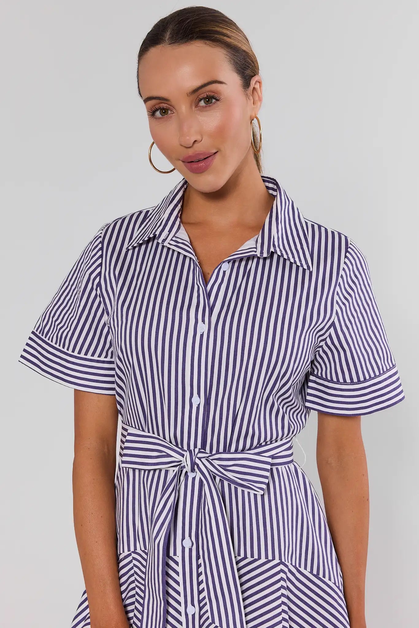 A woman wearing a short-sleeve collared shirt dress with vertical navy and white stripes, featuring a tie waist belt, photographed against a light gray studio background close up