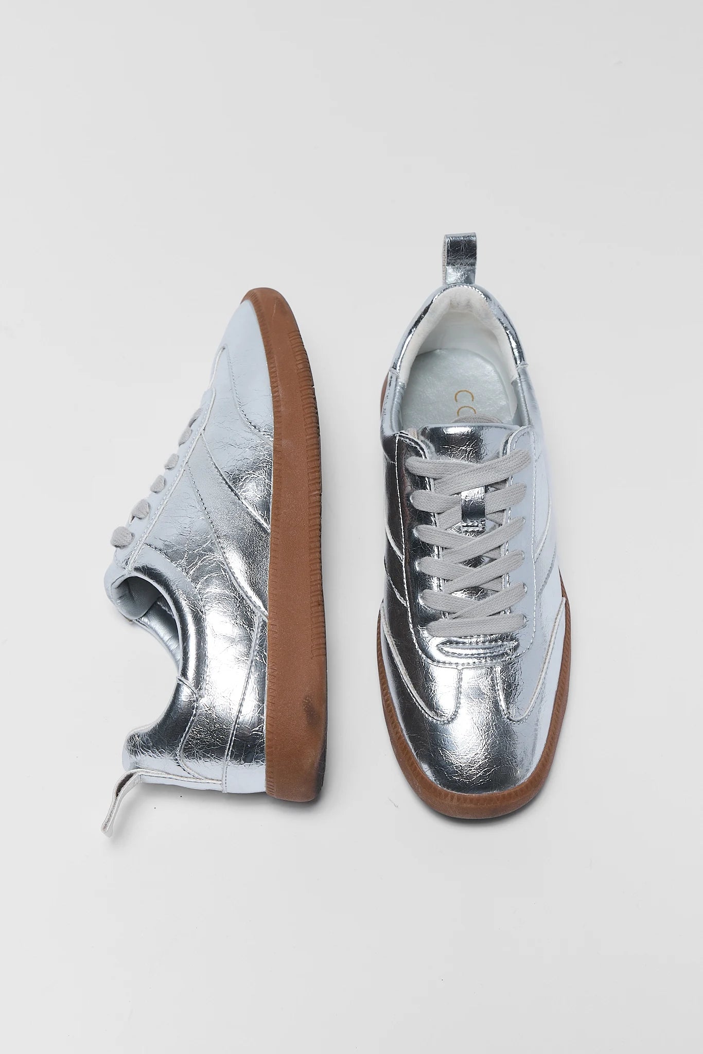 A pair of metallic silver low-top sneakers with white laces and brown rubber soles, shown from side and top-down angles on a light gray background.