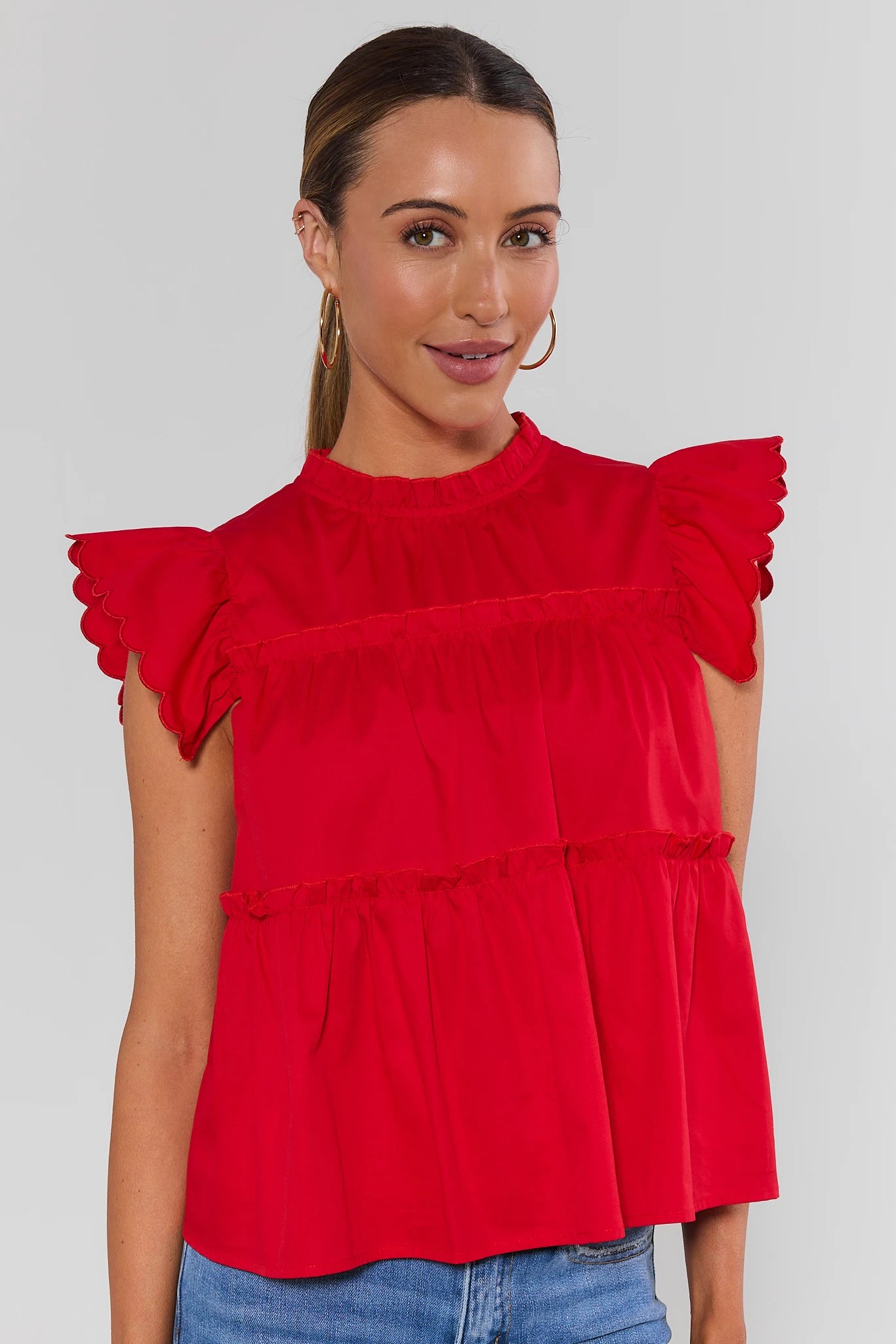 A woman modeling a bright red ruffled short-sleeve top with a high neckline, against a light gray background.