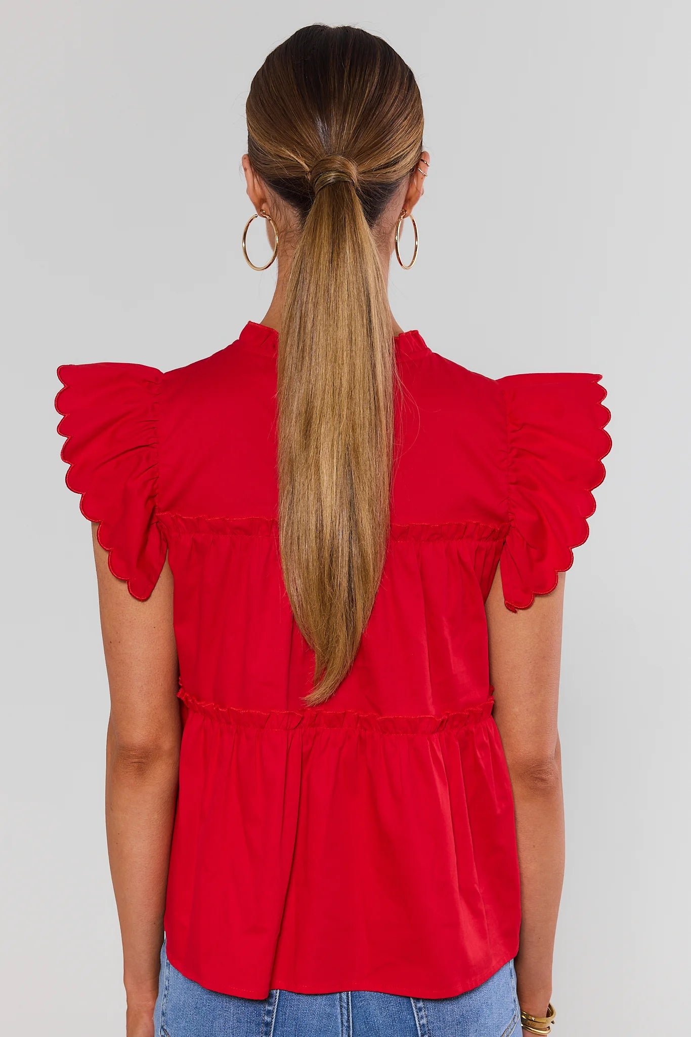 A woman viewed from the back wearing a bright red short-sleeve blouse with ruffled scalloped sleeves and a tiered waist, paired with blue jeans against a light gray studio background.