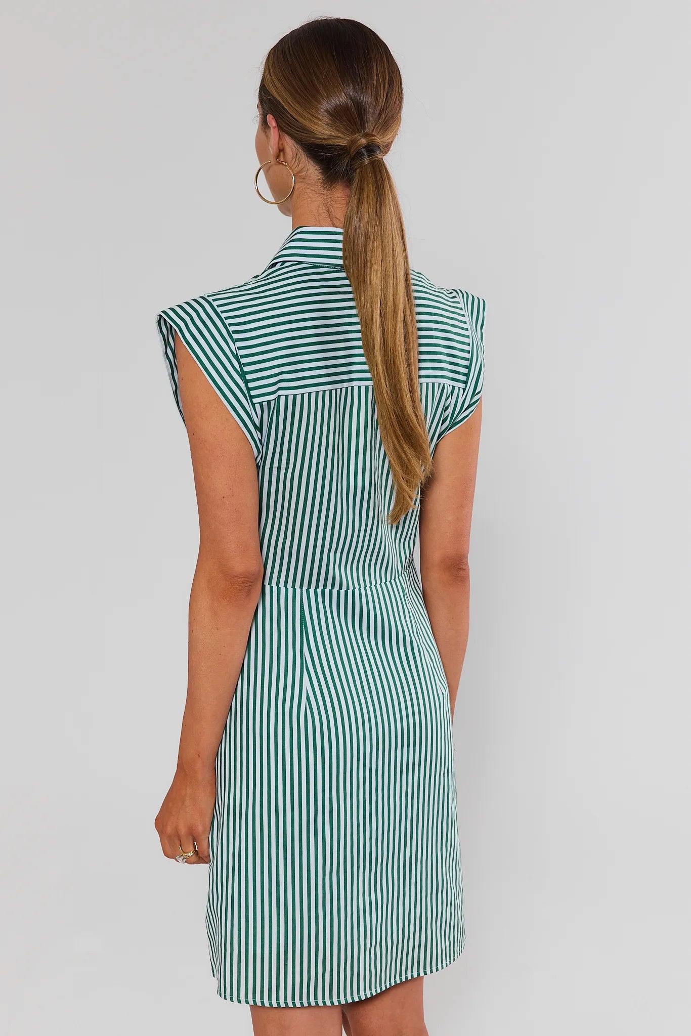 A woman shown from the back wearing a sleeveless collared dress with vertical green and white stripes, photographed against a plain light gray studio background.