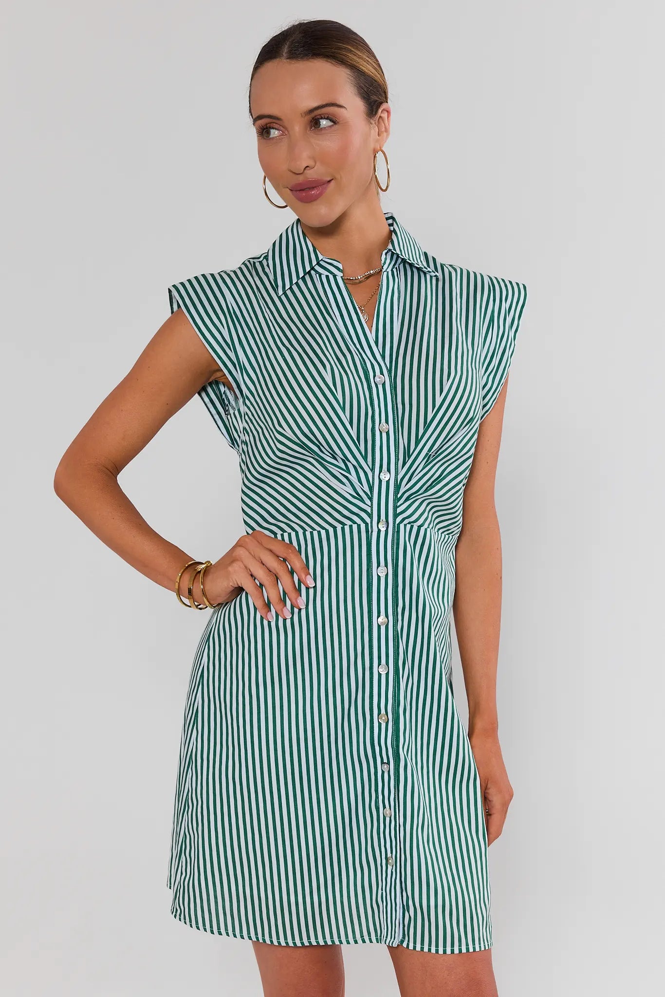 A woman modeling a sleeveless collared shirt dress with vertical green and white stripes, standing against a light gray studio background.