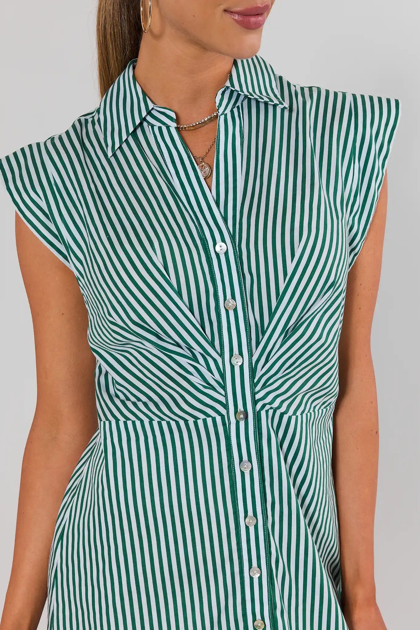 A close-up of a woman wearing a sleeveless collared button-front shirt dress with vertical green and white stripes against a light gray background.