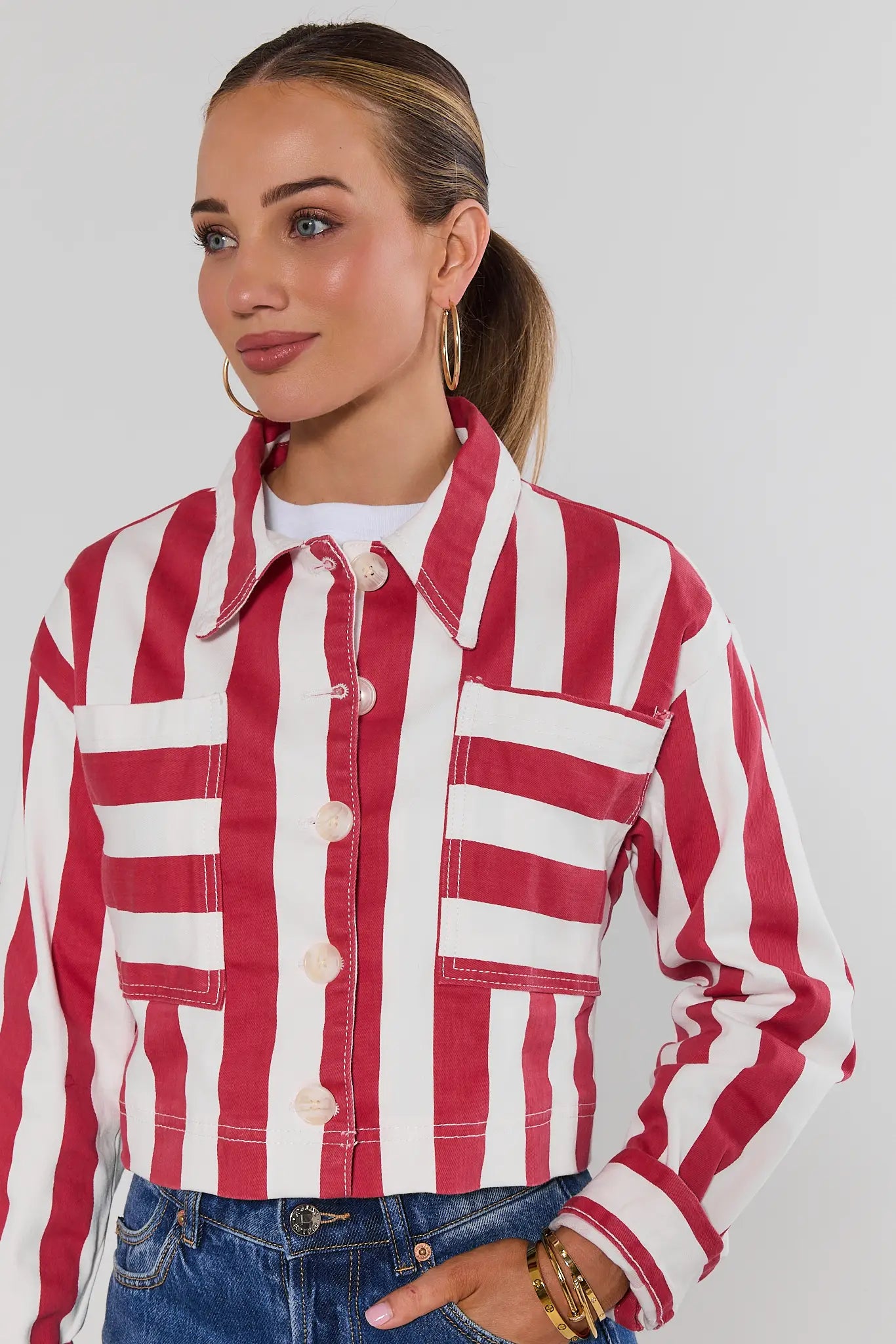 A woman wearing a red and white vertically striped cropped denim jacket over a white t-shirt and blue jeans, photographed against a light gray studio background.