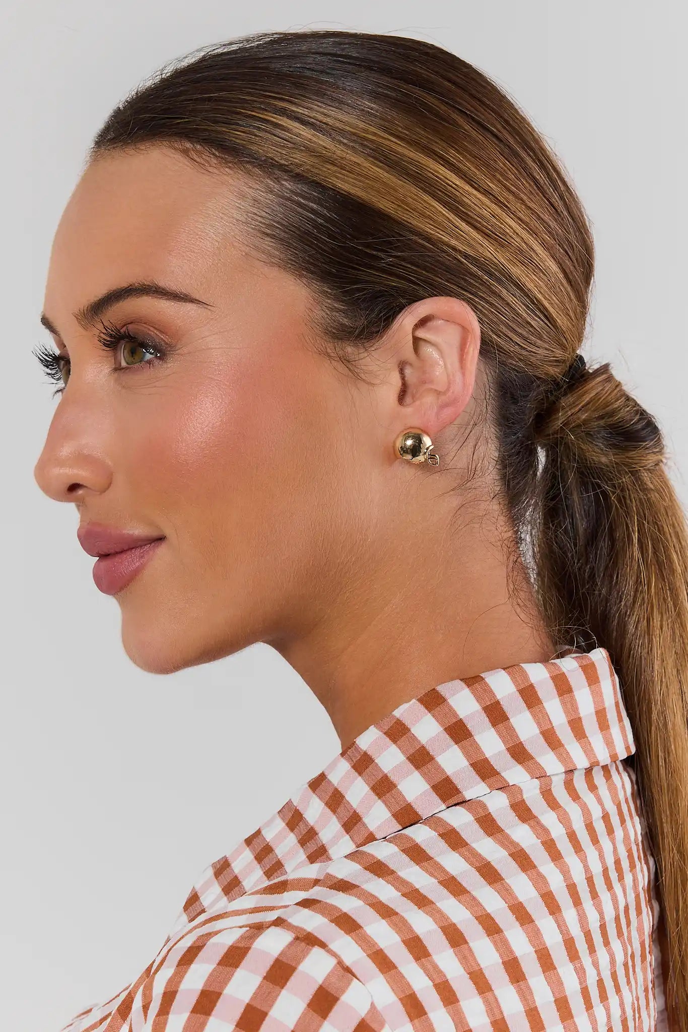 A close-up side profile of a woman with brown hair in a ponytail, wearing a brown and white gingham shirt and a small gold football helmet earring, against a light gray background.