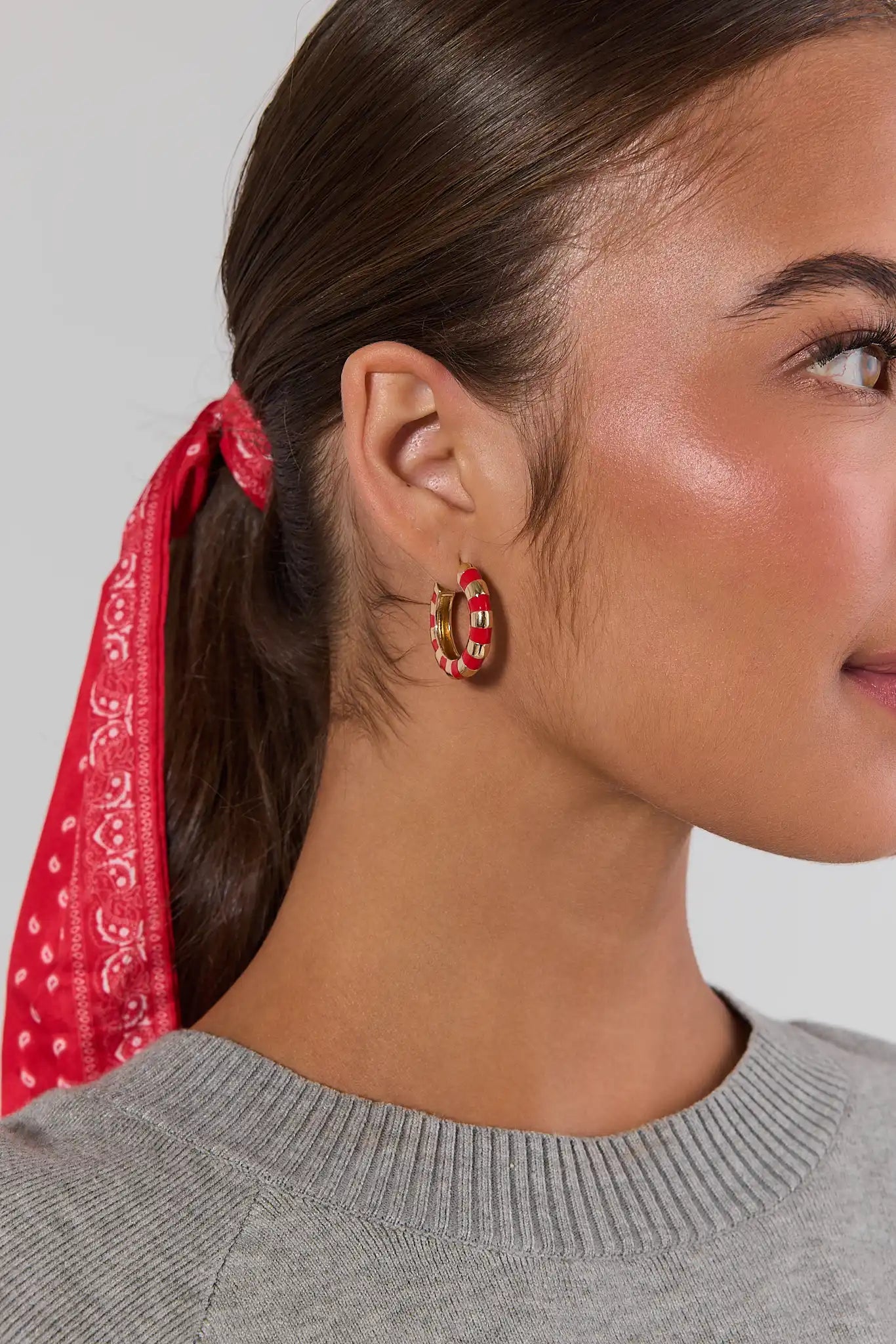 A close-up side profile of a woman wearing a pair of chunky gold hoop earrings with red and white patterned inlay, with her brown hair tied back with a red paisley bandana.
