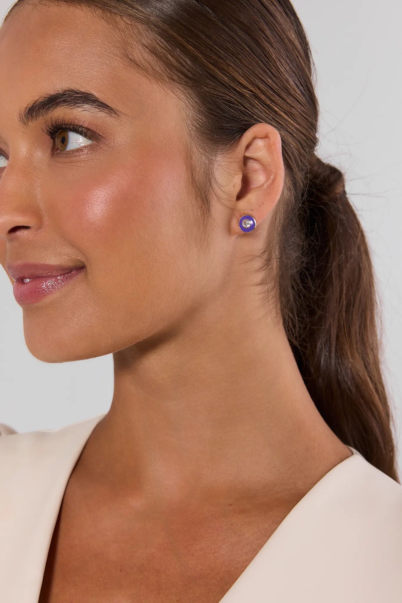 A close-up side profile of a woman with brown hair in a ponytail, wearing a small round purple stud earring, against a light gray background.