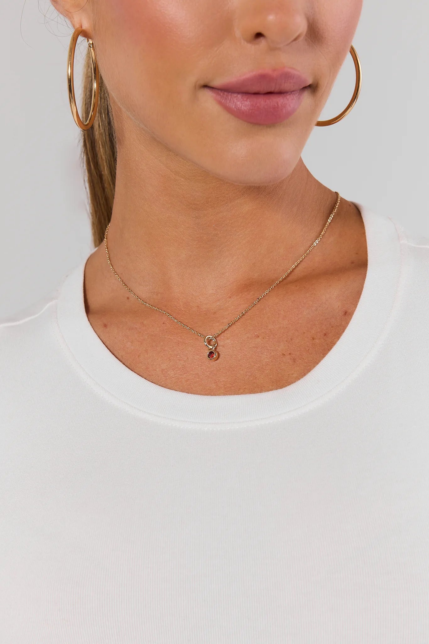 A close-up of a model wearing a delicate gold chain necklace with a small red charm pendant, styled with gold hoop earrings and a white crew-neck top.