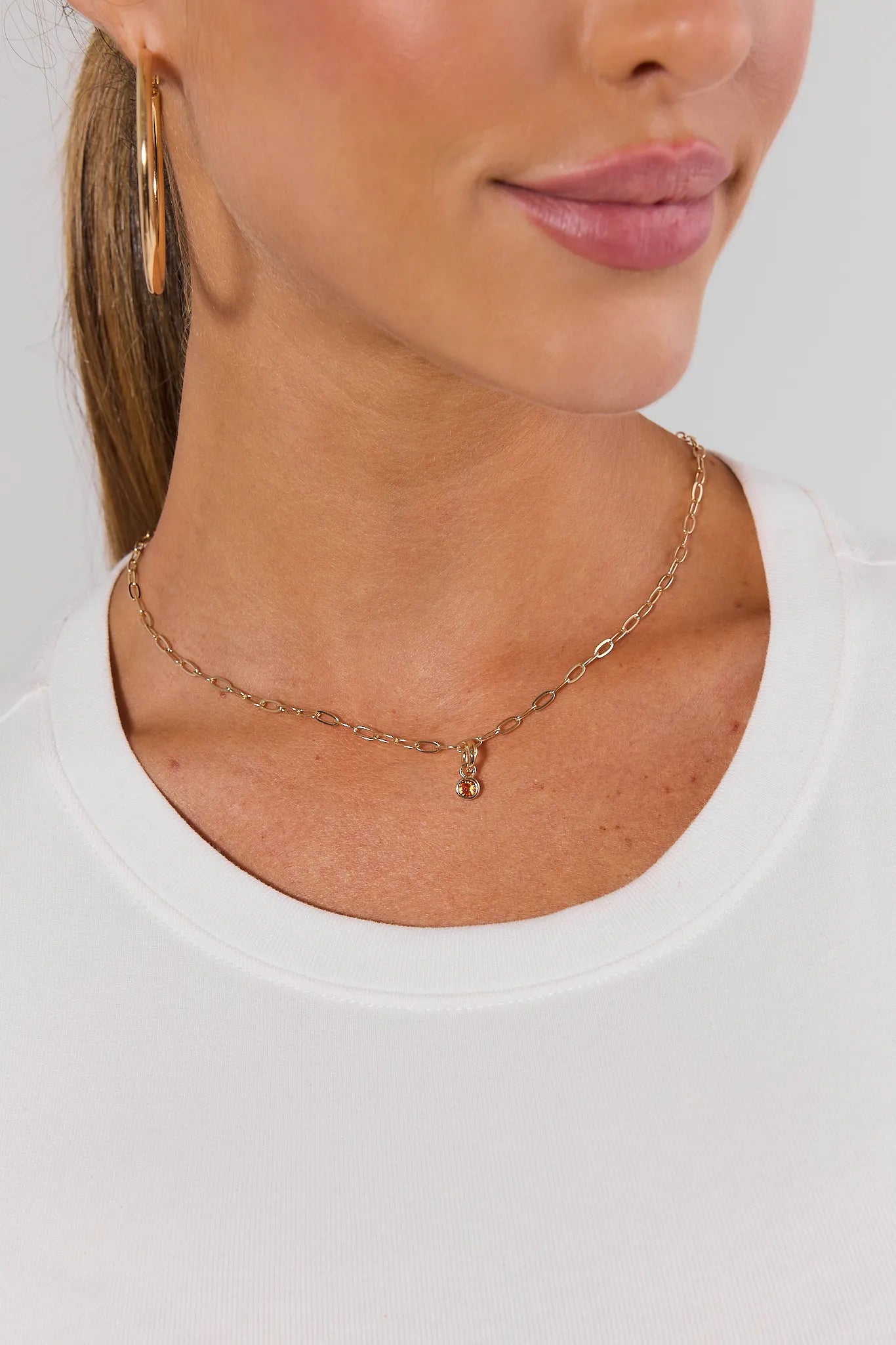A woman wearing a necklace with a small gold charm featuring a dangling oval amber gemstone, centered on a plain light gray background.