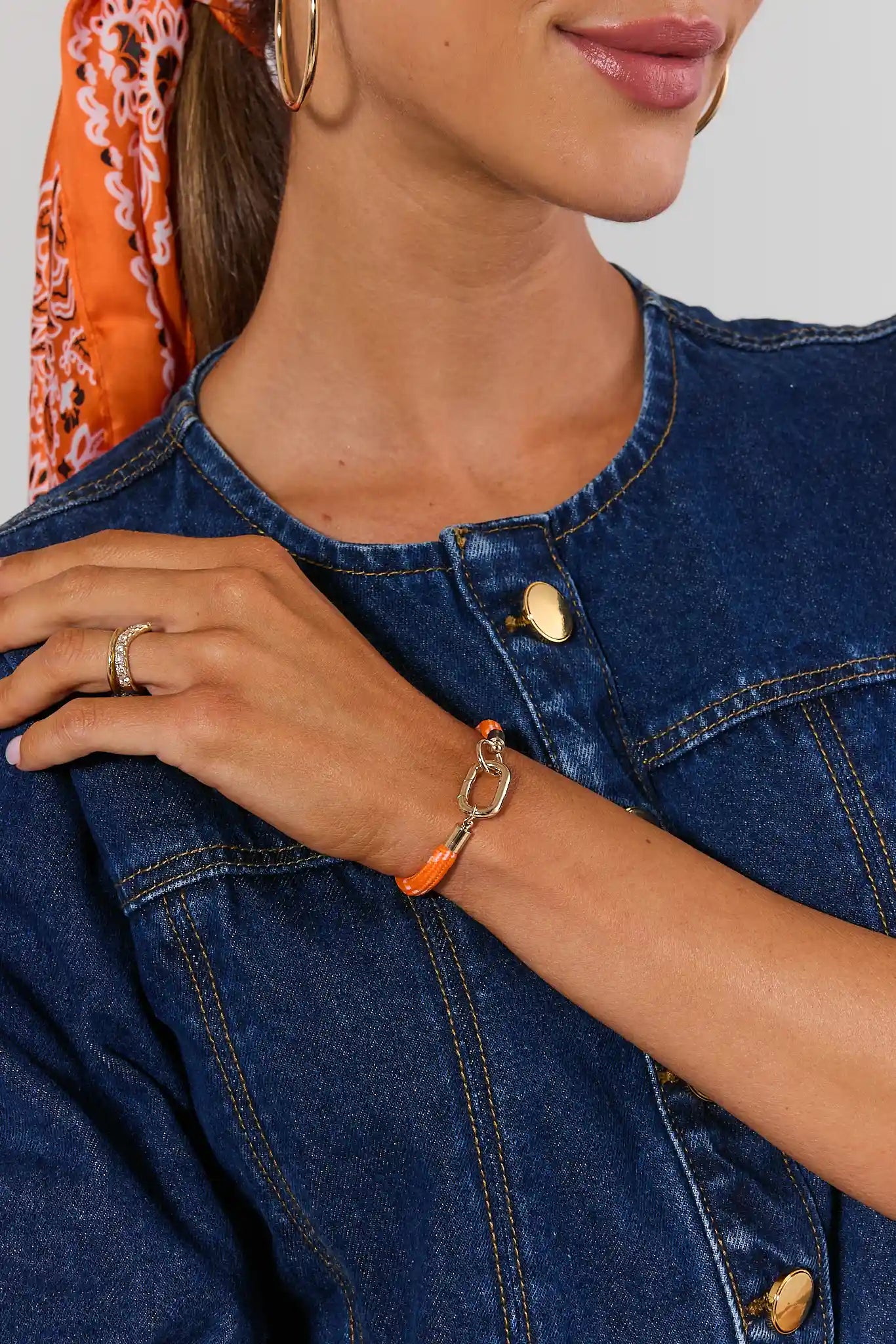 A close-up of a woman wearing a gold-tone chain bracelet with a bright orange wrap detail on her wrist, paired with a denim jacket and hoop earrings.