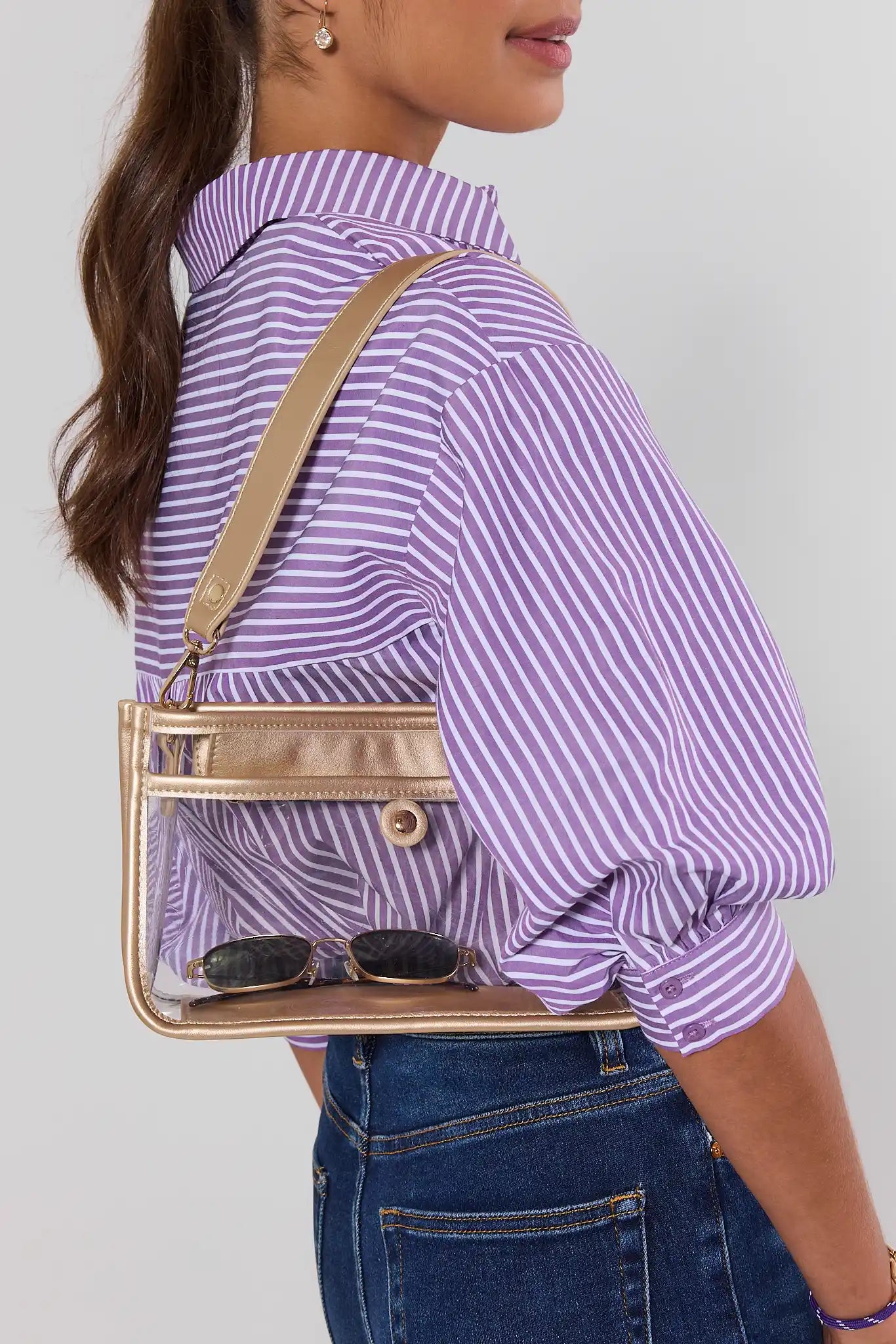 A woman wearing a lavender and white striped shirt and blue jeans carries a clear rectangular shoulder bag with a metallic gold frame and strap. The bag has sunglasses hanging from the front pocket.