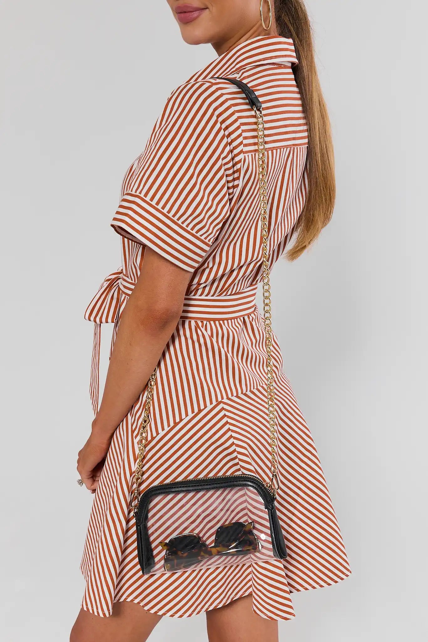 A woman wearing a short-sleeve, collared dress with vertical red and white stripes, shown from a side-back angle. She is carrying a small clear pouch crossbody bag with a gold chain strap and black trim.