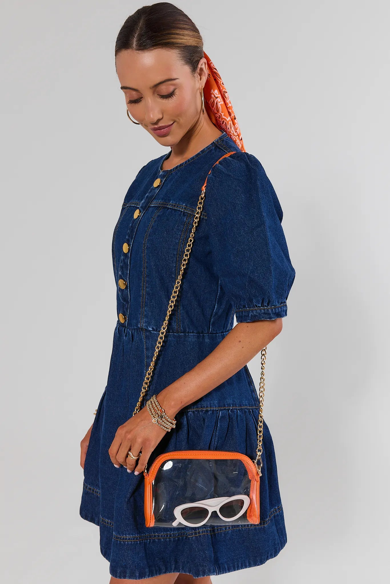 A woman wearing a blue denim dress with gold buttons and puff sleeves, carrying a clear crossbody bag with an orange trim and chain strap. She is posed against a light gray studio background.
