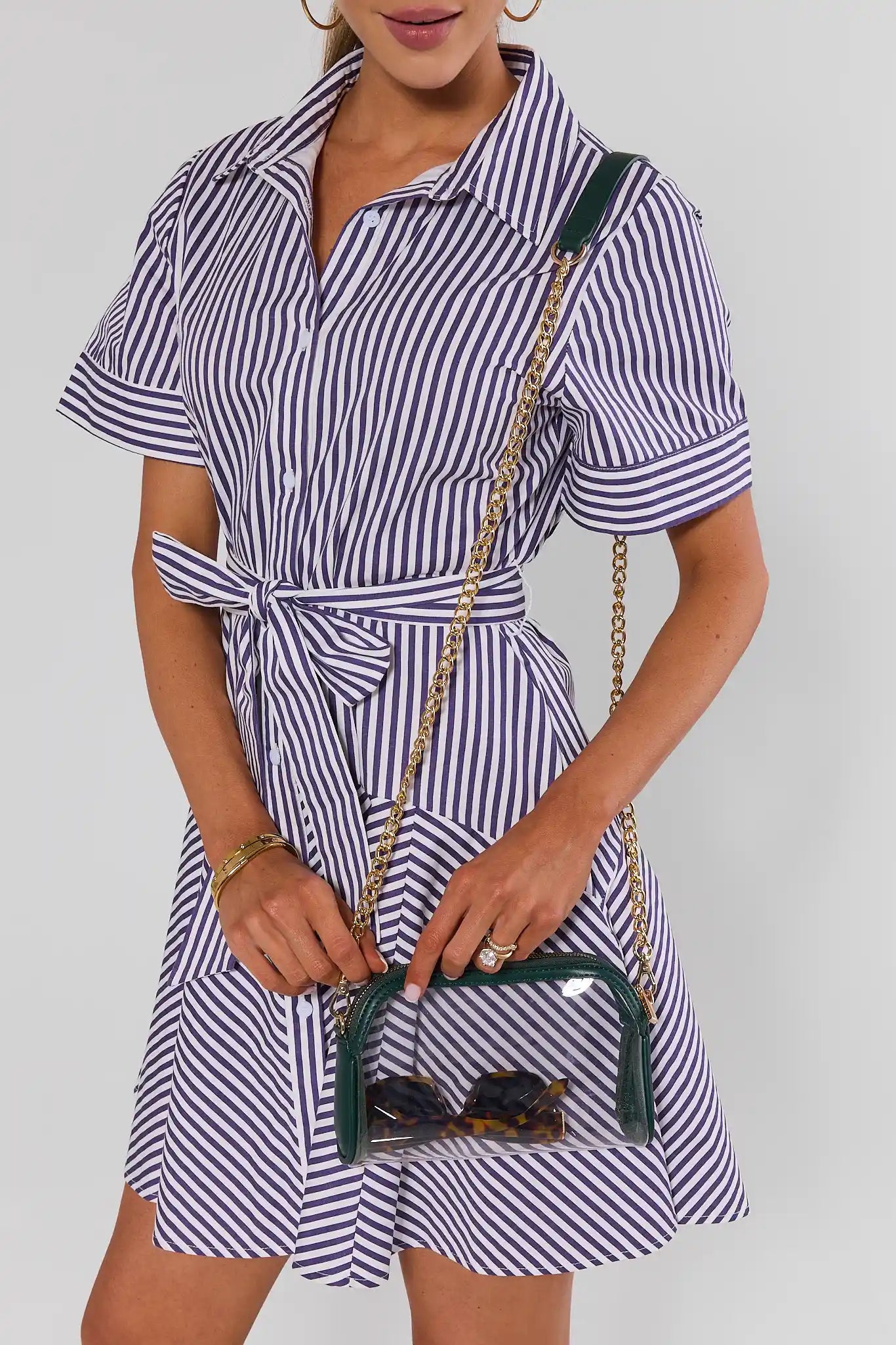 A woman wearing a short-sleeve button-up shirt dress with vertical navy and white stripes, holding a clear pouch handbag with a green frame and gold chain strap.