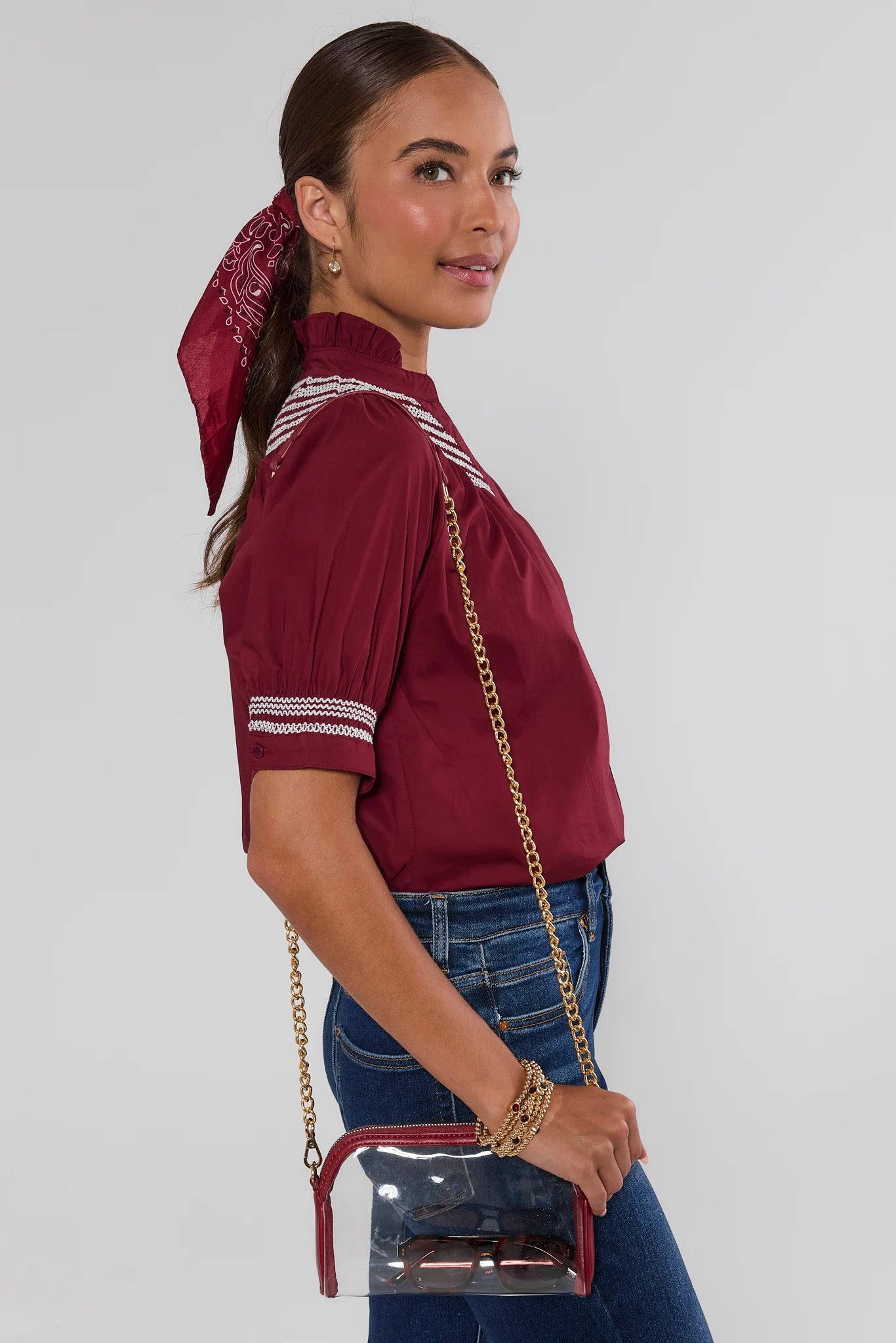 A woman wearing a burgundy short-sleeve blouse with white stripe details on the collar and sleeves, paired with blue jeans and a small clear crossbody bag with red trim and a gold chain strap.