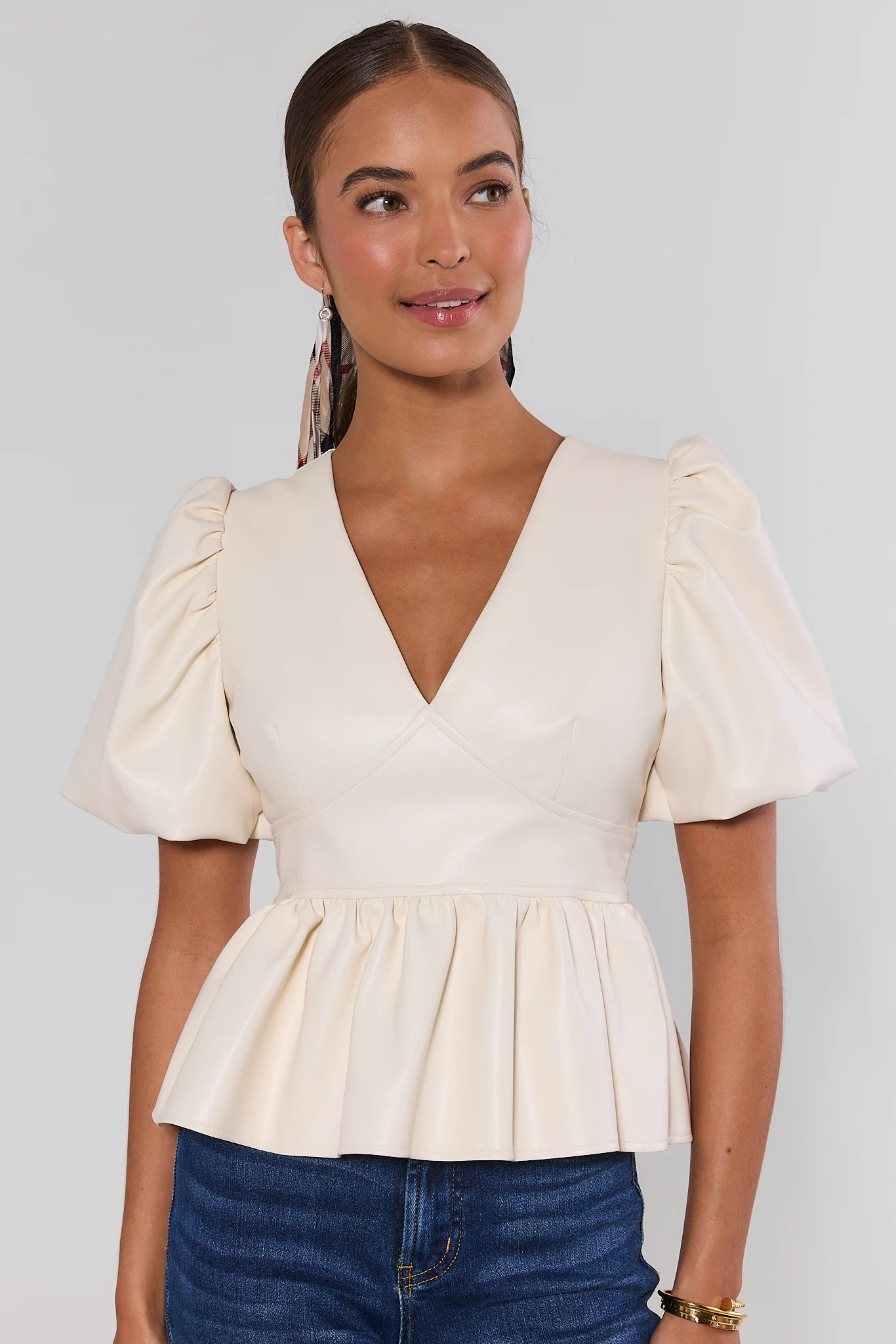 A woman wearing a cream-colored peplum top with puff sleeves and a deep V-neck, paired with blue jeans, against a light gray studio background.