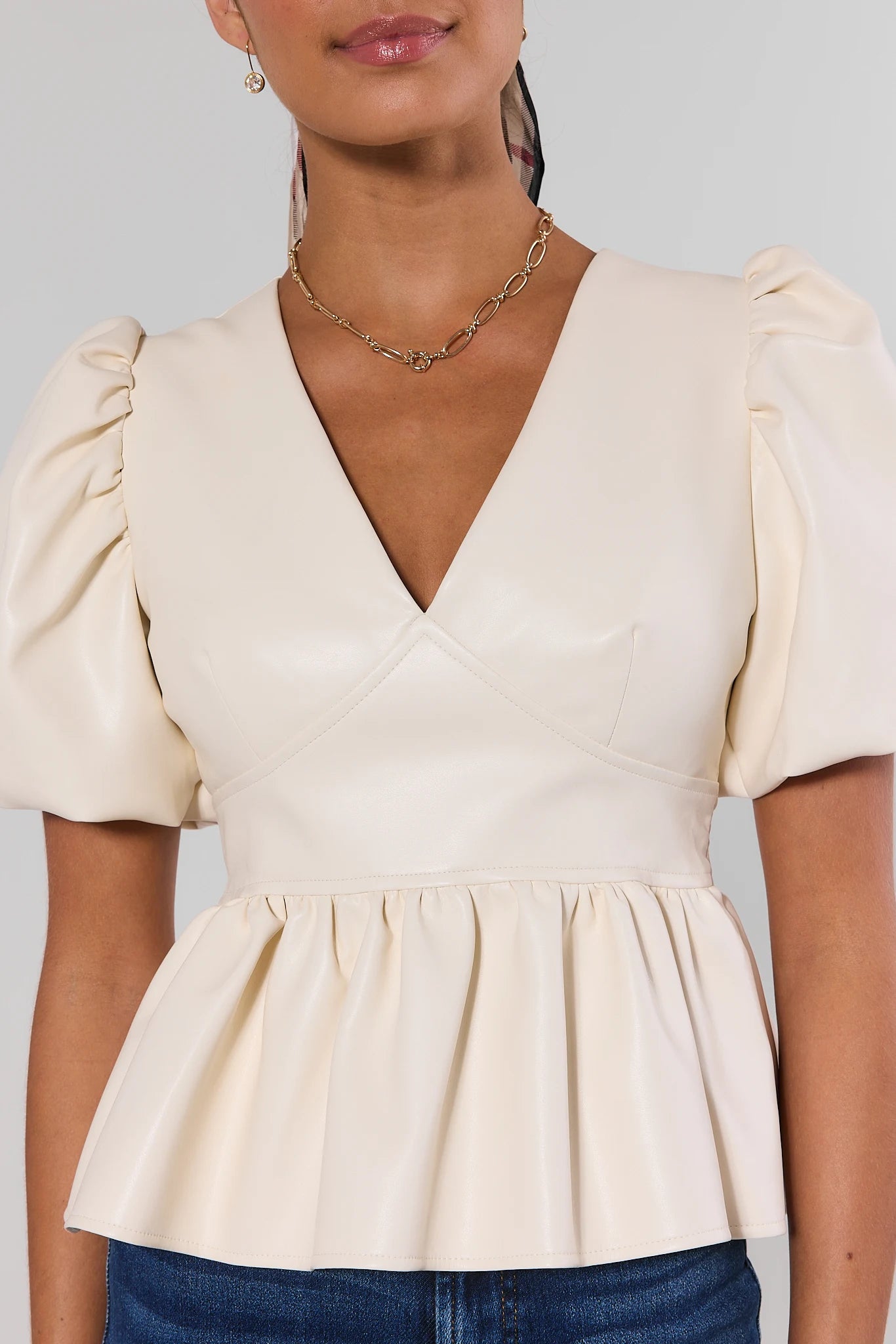 A woman wearing a cream-colored faux leather peplum top with puff sleeves and a wrap-style V-neckline, paired with blue jeans close up