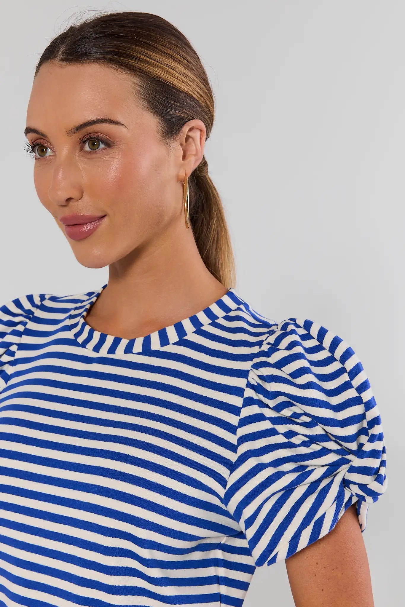 Close up a woman wearing a blue and white horizontally striped short-sleeve top with puffed sleeves, shown from the chest up against a light gray studio background.