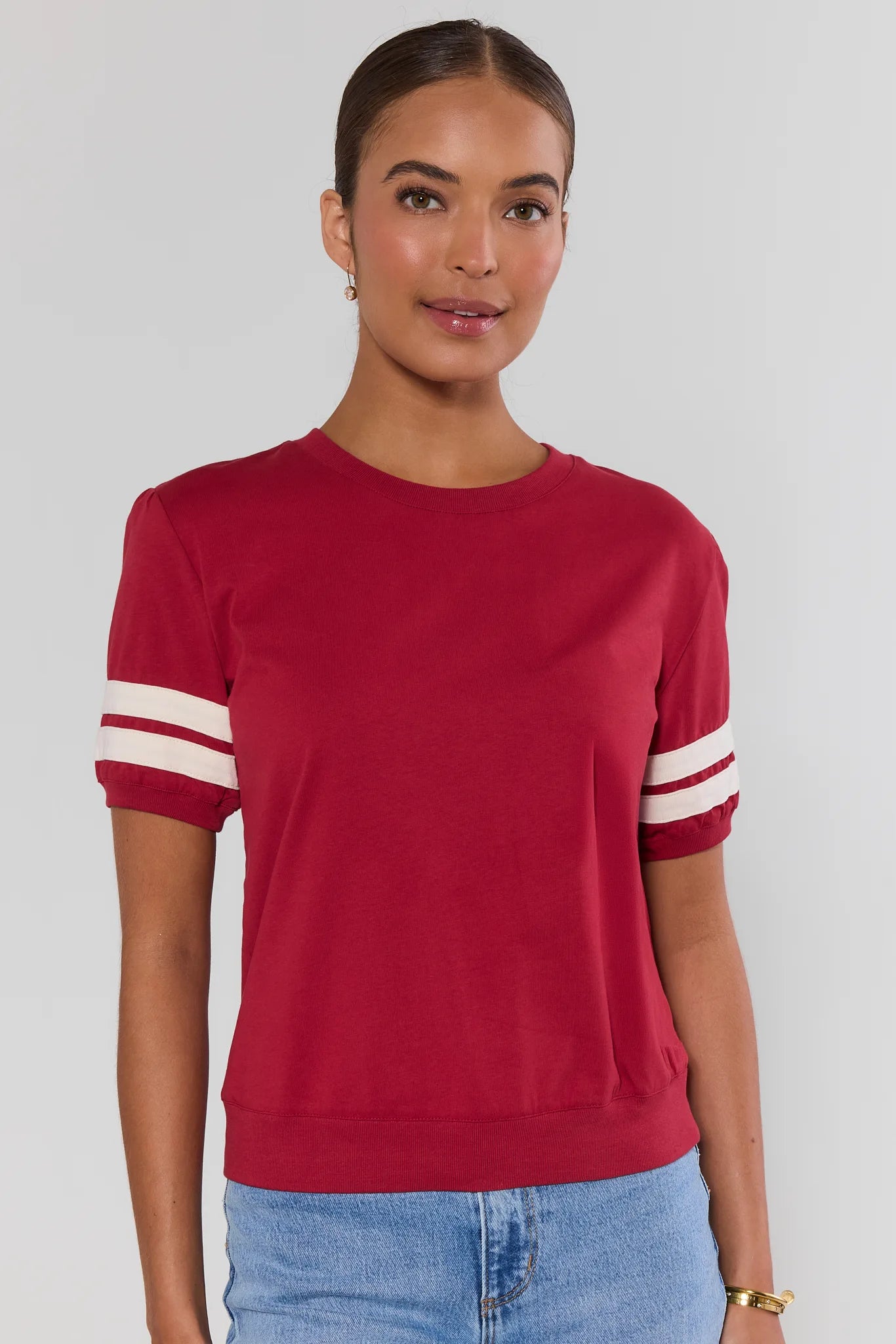 A woman wearing a short-sleeve crewneck top in deep red with white stripes on the sleeves, paired with light blue jeans, against a plain light gray background.