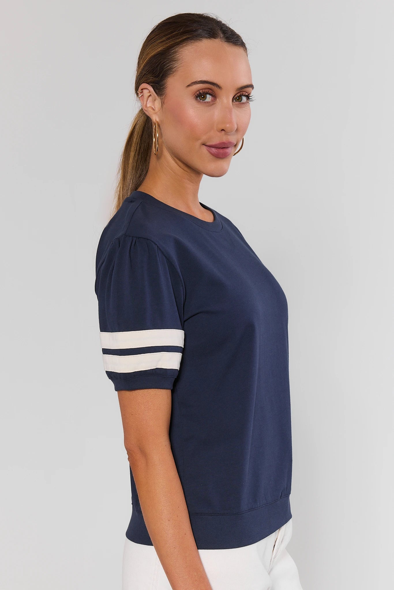 A woman from the side modeling a navy blue short-sleeve t-shirt with white stripes on the sleeves, paired with white wide-leg pants, against a light gray studio background.