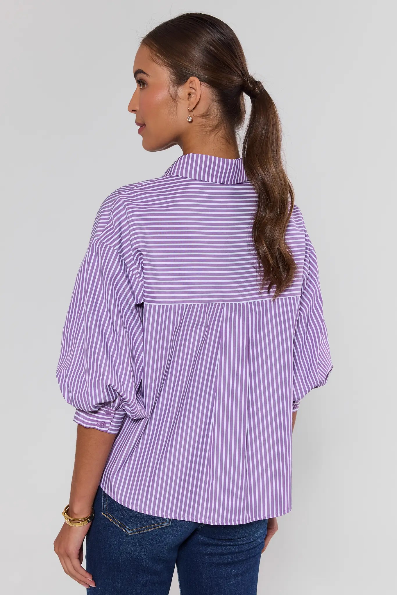 A woman shown from the back wearing a lavender and white vertically striped blouse with puff sleeves and blue jeans, photographed against a plain light gray studio background.