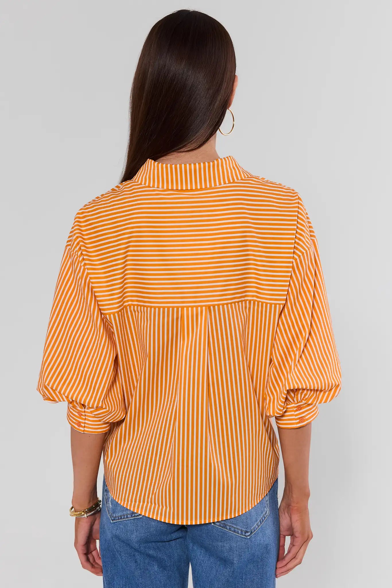 A woman viewed from the back wearing a yellow and white vertically striped blouse with puffed three-quarter sleeves and blue jeans, against a light gray studio background.