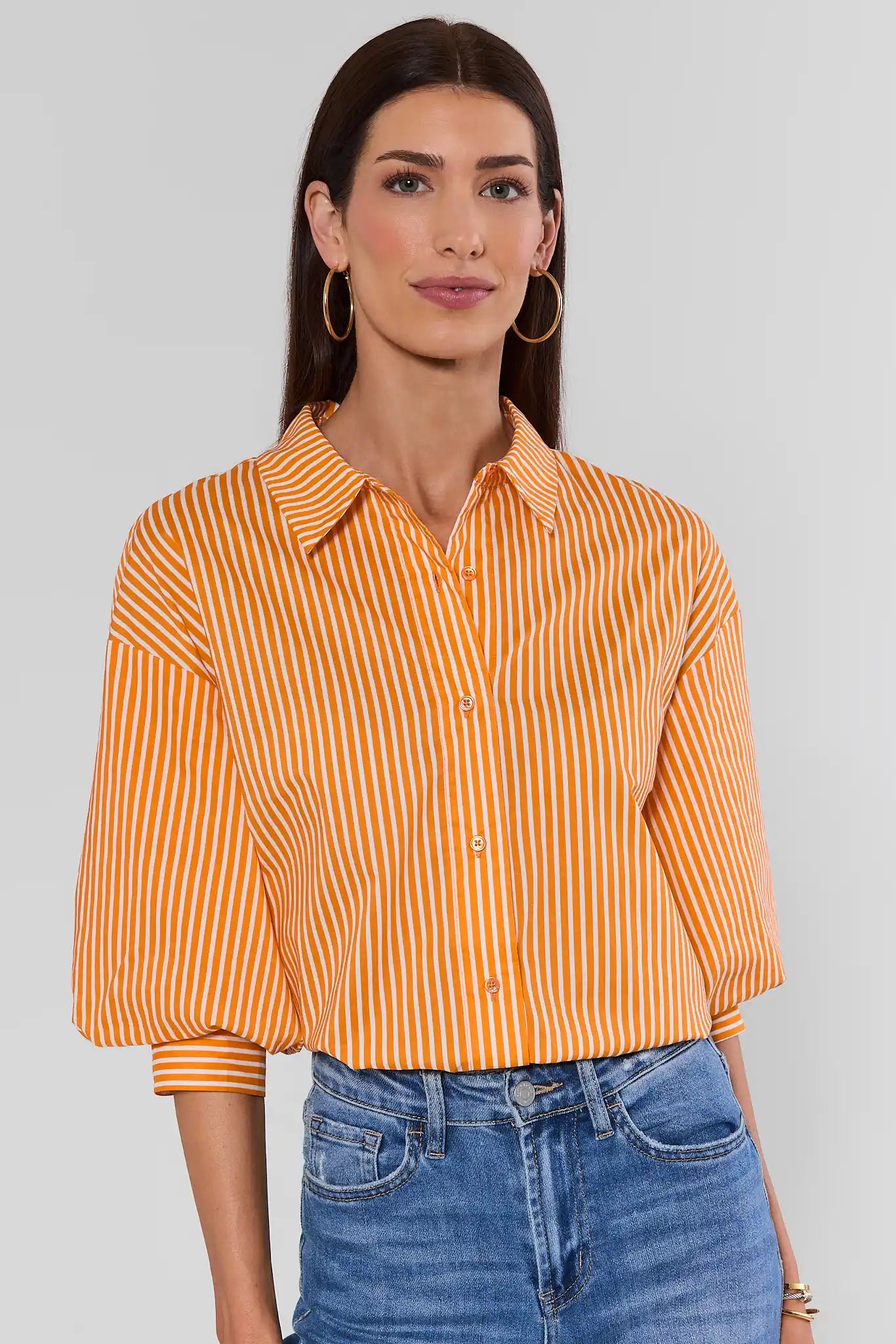 A woman wearing a cropped orange and white vertically striped button-up shirt with three-quarter sleeves, paired with blue jeans, against a light gray studio background.