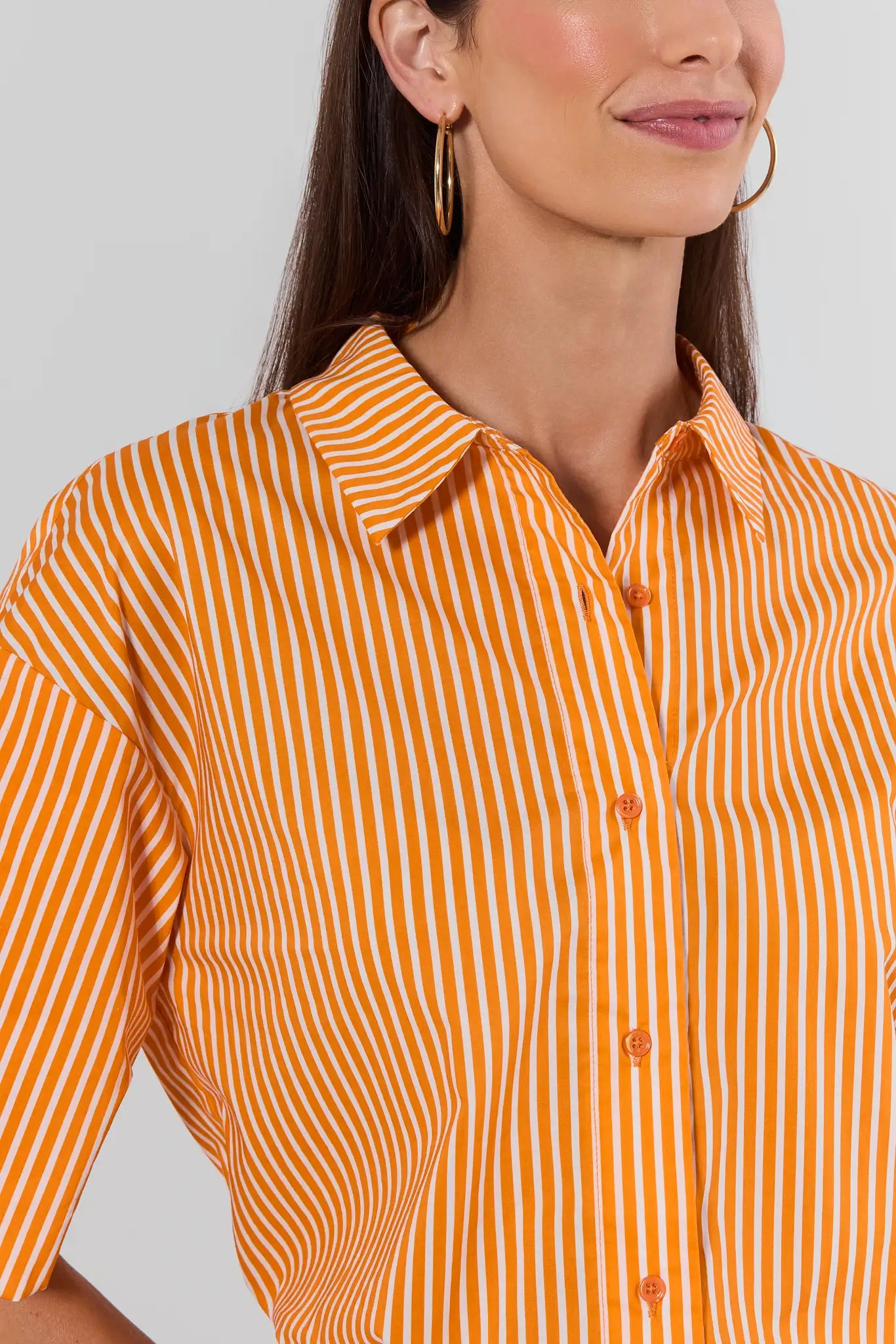 A close-up of a woman wearing a short-sleeve button-up shirt with vertical orange and white stripes, paired with gold hoop earrings, against a light gray background.