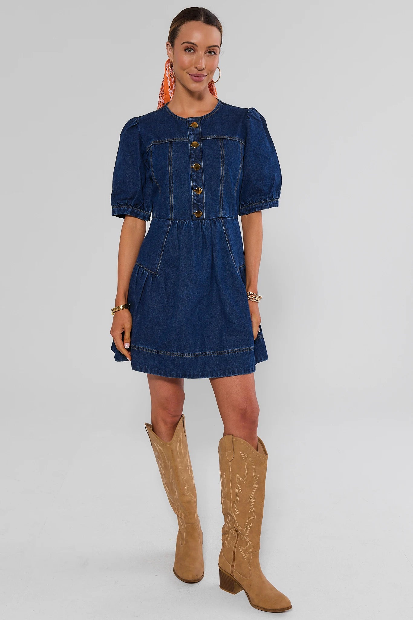 A woman modeling a short blue denim dress with puff sleeves and a button-front bodice, paired with tan cowboy boots, against a light gray studio background.