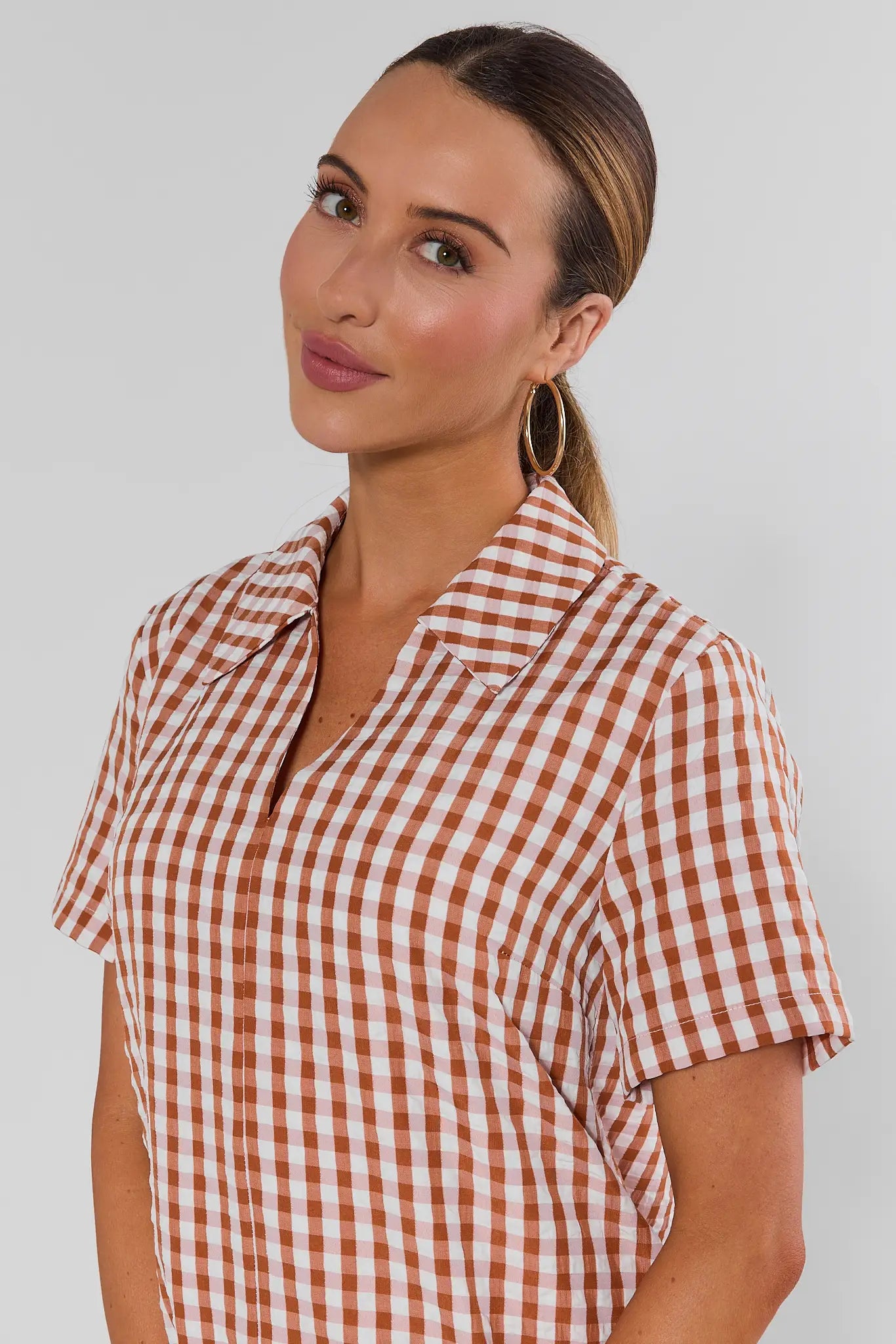 A woman wearing a short-sleeve collared button-up shirt with a rust orange and white gingham check pattern, photographed against a light studio background.