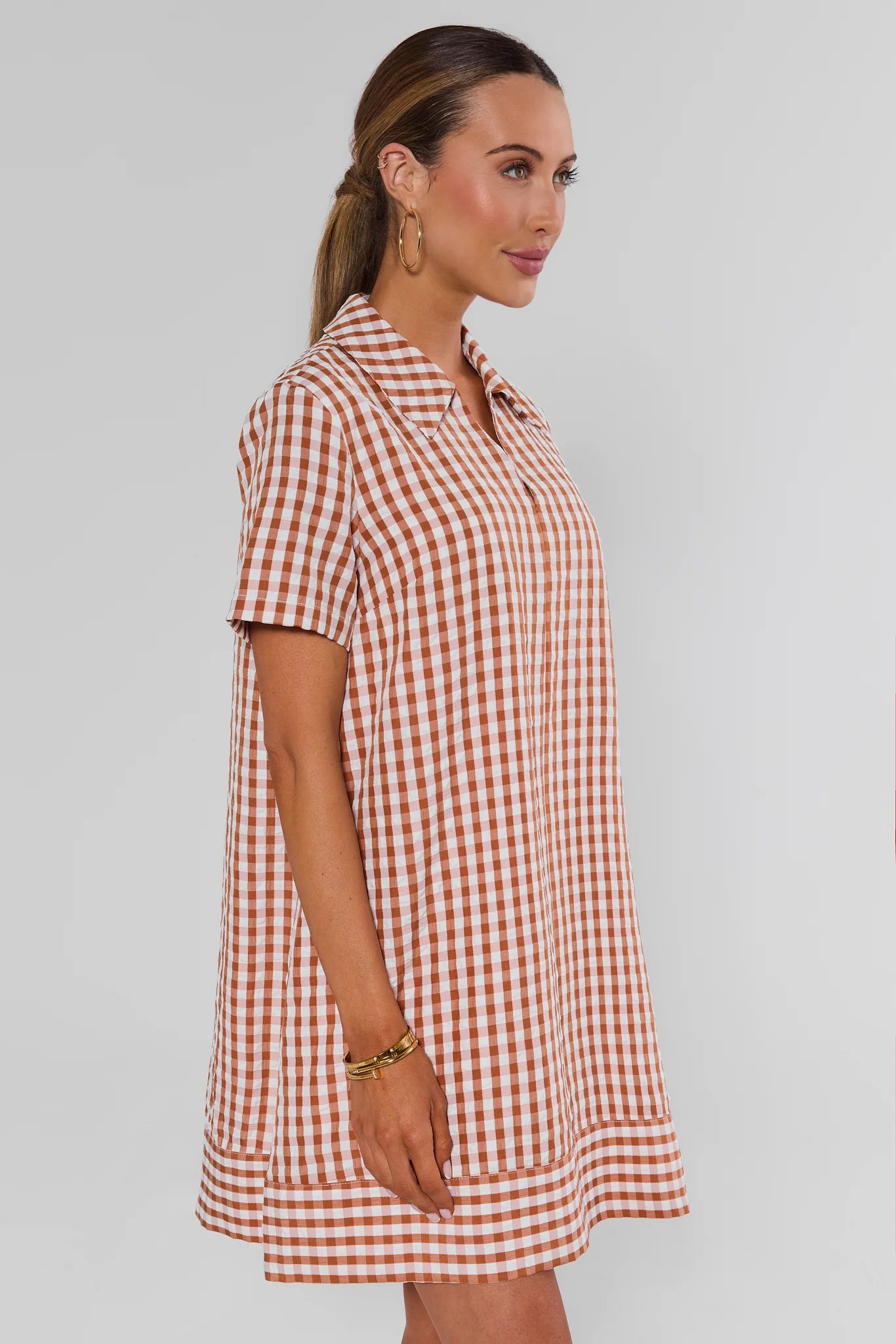 A woman wearing a short-sleeve collared tunic dress with a red and white gingham check pattern, shown from a side profile against a light gray studio background.