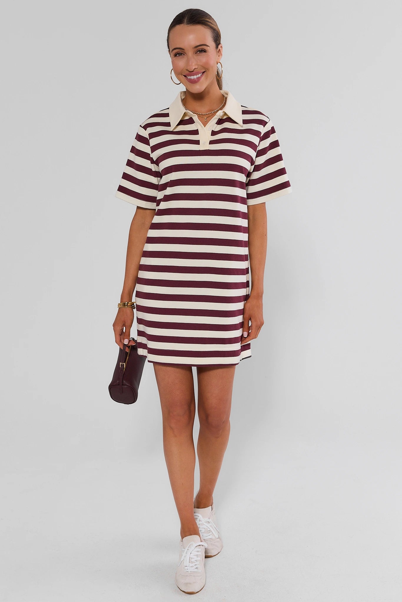 A woman walking wearing a short-sleeve polo dress with horizontal burgundy and cream stripes, featuring a solid cream collar and placket, set against a plain light gray studio background.
