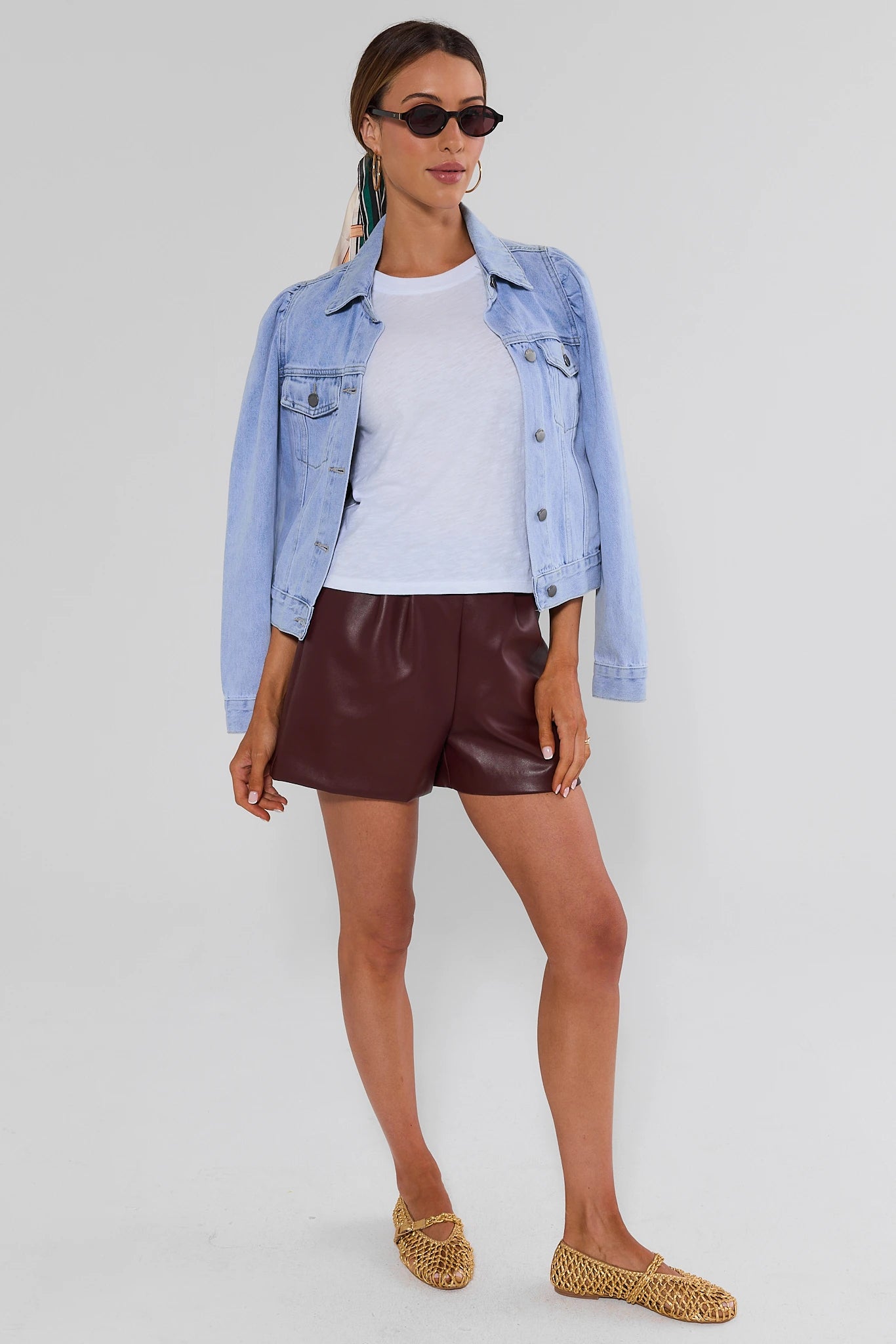 A model wearing a jean jacket and high-waisted, shiny burgundy faux-leather shorts and woven tan flat shoes, shown from the waist down against a light studio background.