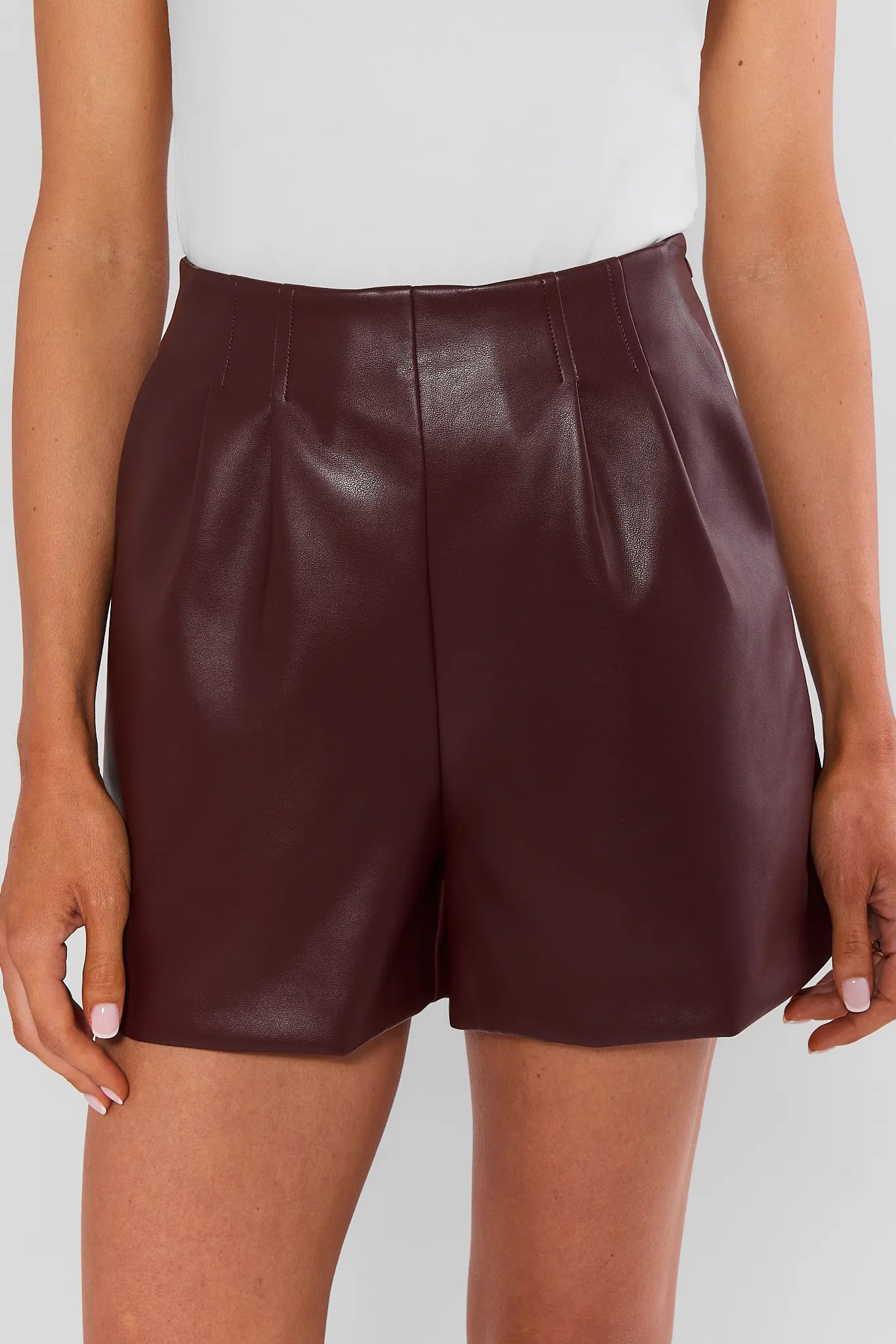 A model wearing high-waisted, shiny burgundy faux-leather shorts, shown from the waist down against a light gray studio background.