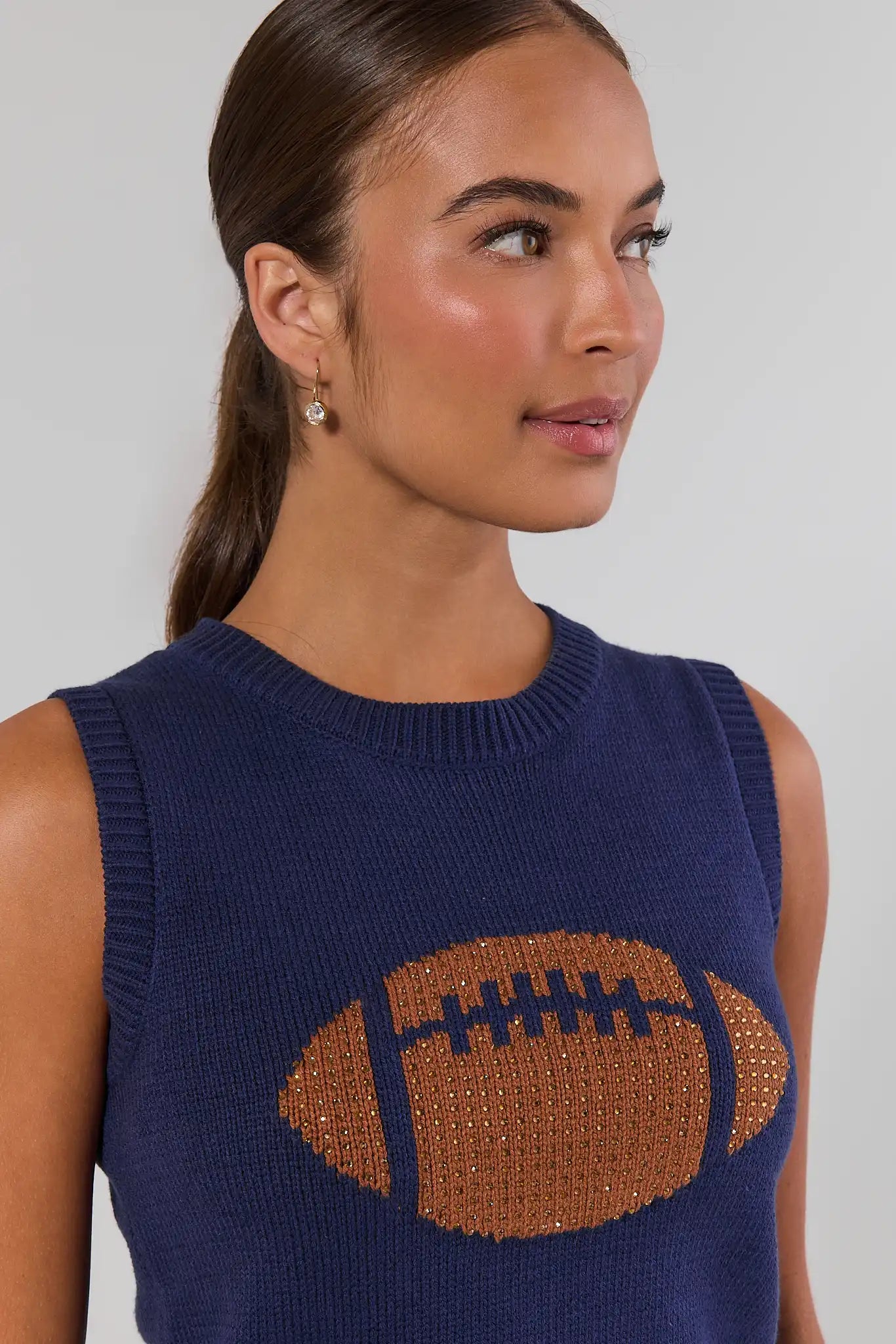 A woman wearing a navy blue sleeveless knit sweater with a large brown football graphic on the front, shown against a light gray studio background close up