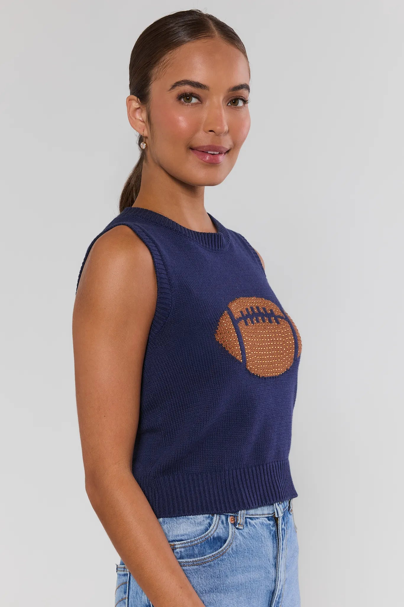 A woman wearing a navy blue sleeveless knit sweater with a large brown football graphic on the front, shown against a light gray studio background turned to the side
