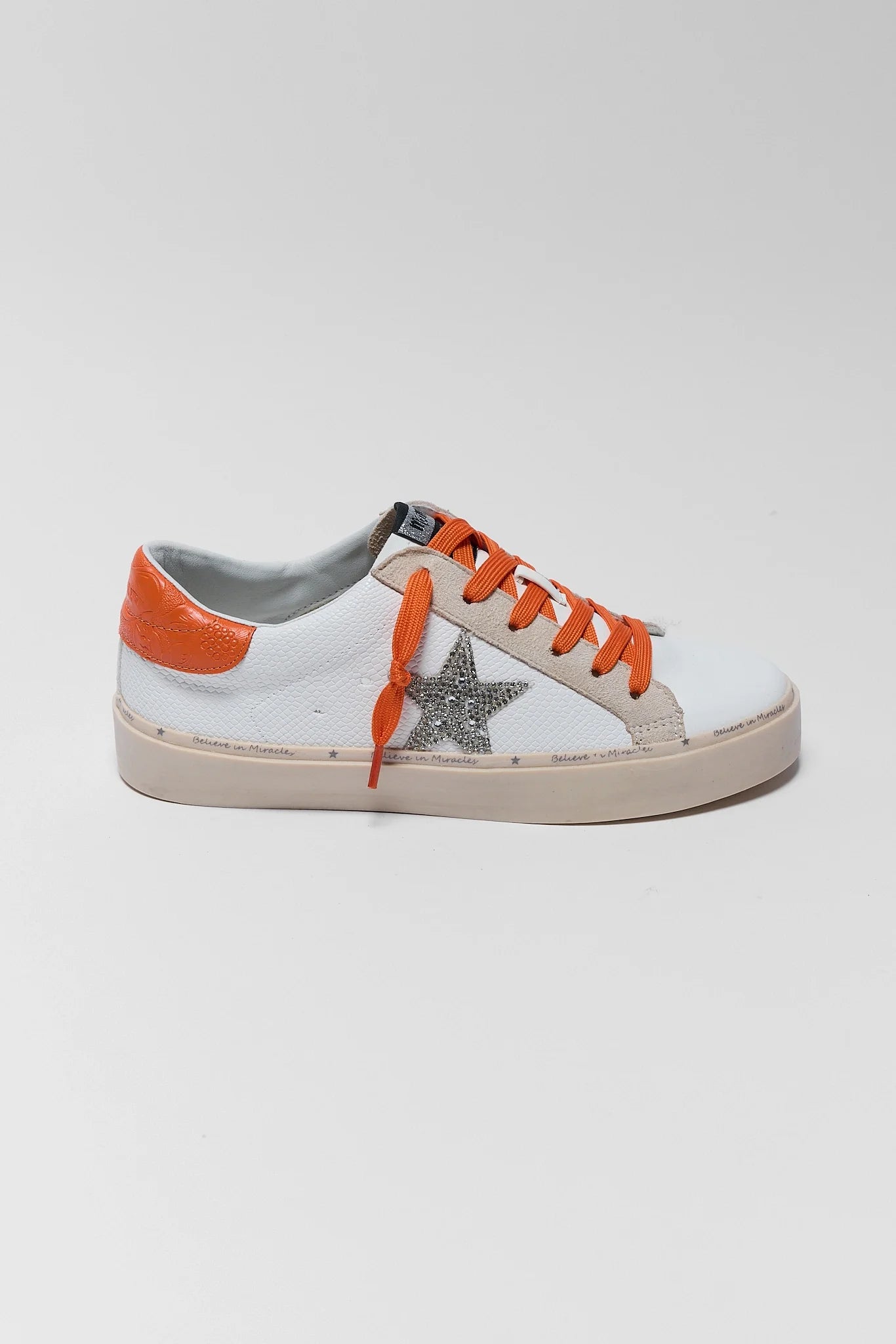 A white leather low-top sneaker with a silver glitter star on the side, orange laces, and an orange heel tab, shown against a light gray background.