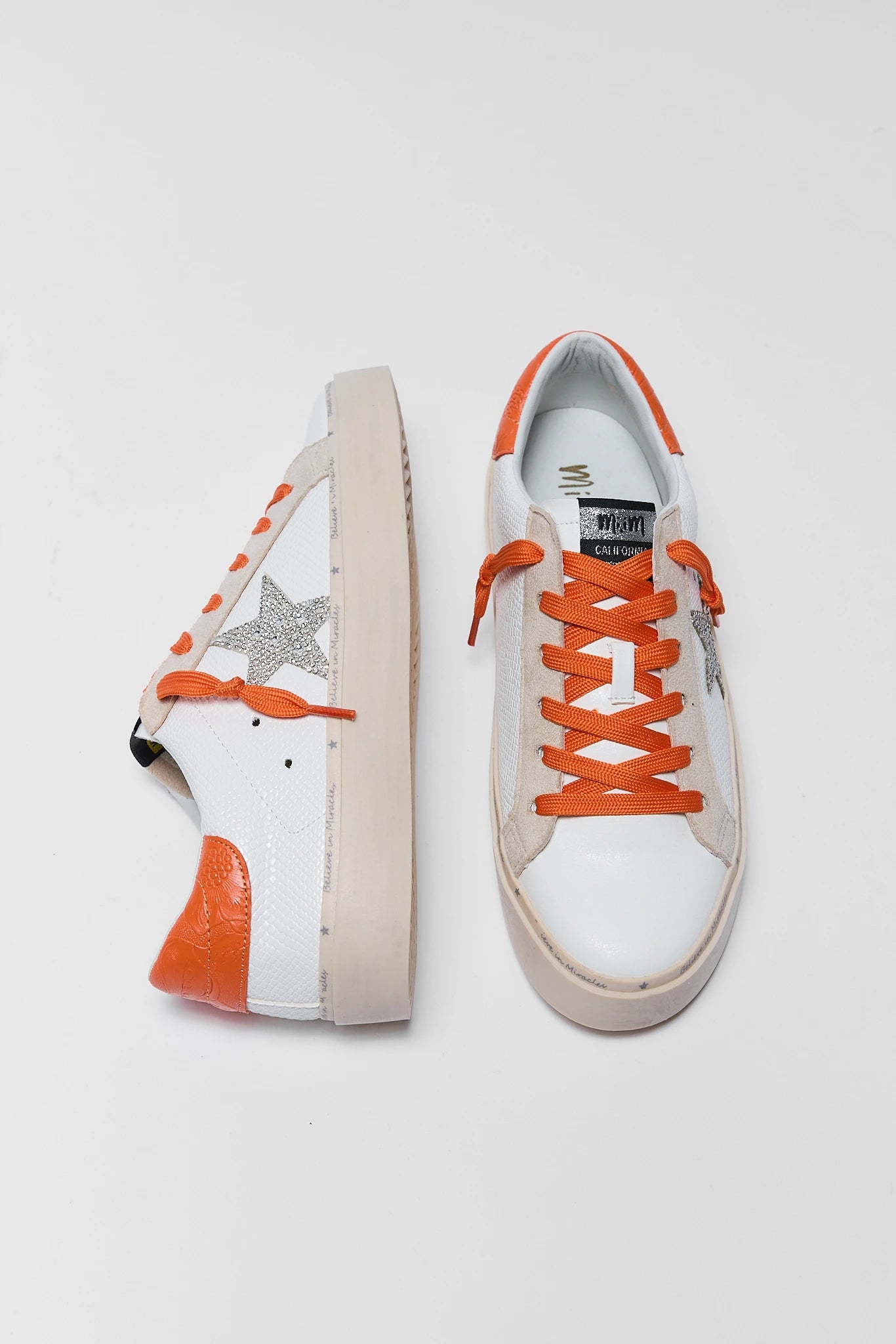 A pair of white leather sneakers with vibrant orange laces, orange heel accents, and a silver glitter star on the side, displayed on a light gray background.