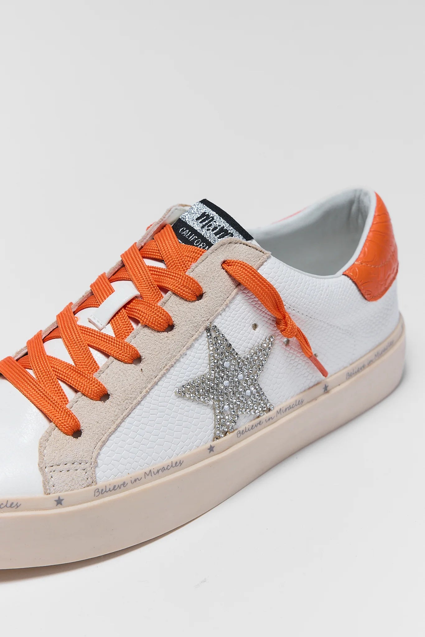 A white leather sneaker with vibrant orange laces, an orange heel tab, and a glittery silver star on the side, shown against a light background.