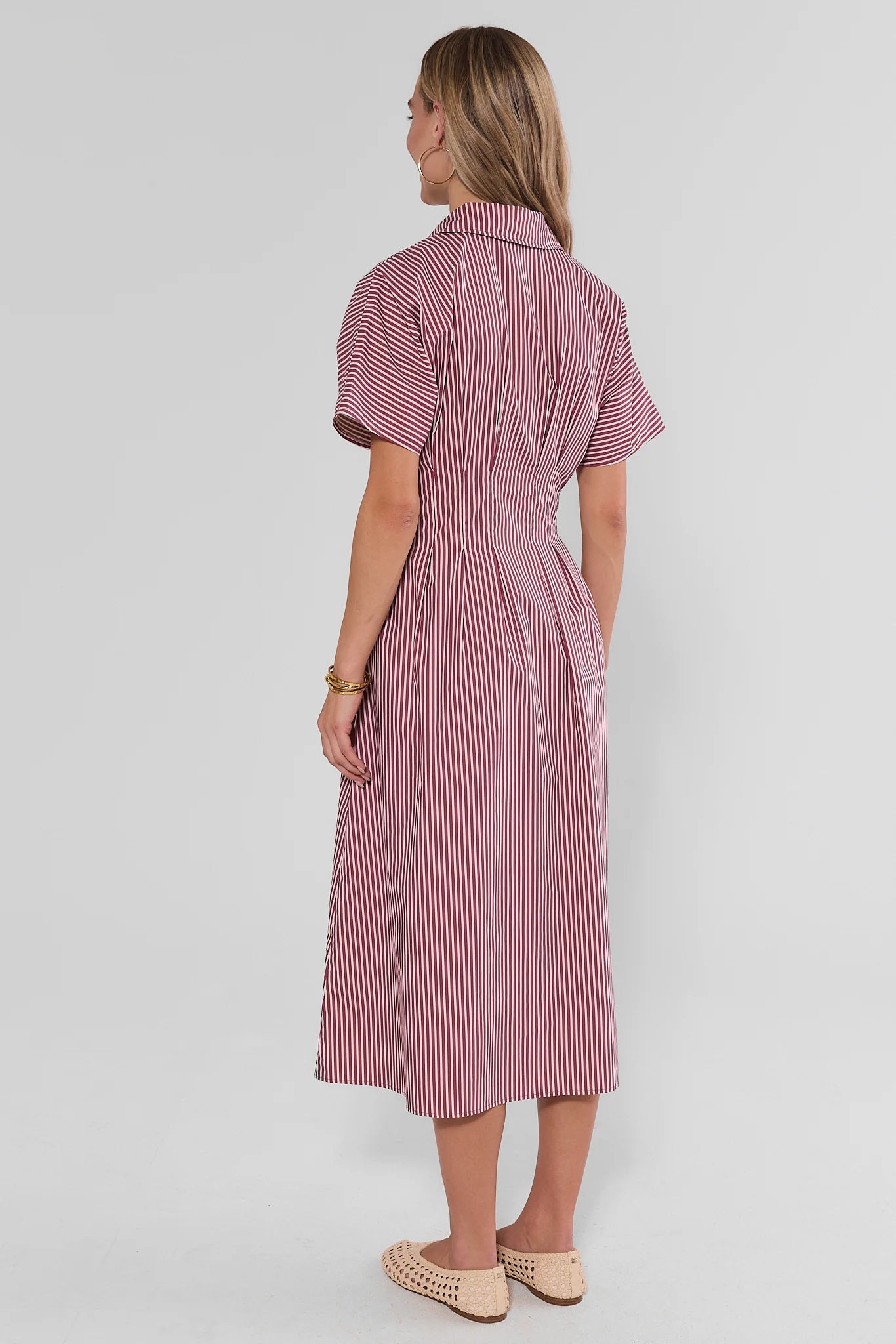 A woman is shown from the back wearing a short-sleeved midi dress with vertical pink and white stripes, paired with beige woven flat shoes, against a light gray studio background.