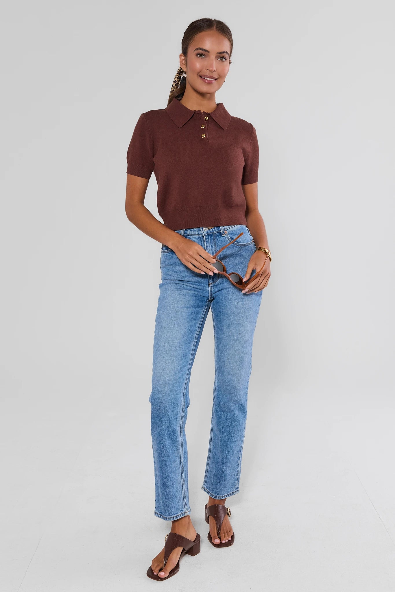 A woman modeling a short-sleeve brown knit polo shirt with light-wash blue jeans and brown sandals, standing against a light gray studio background.