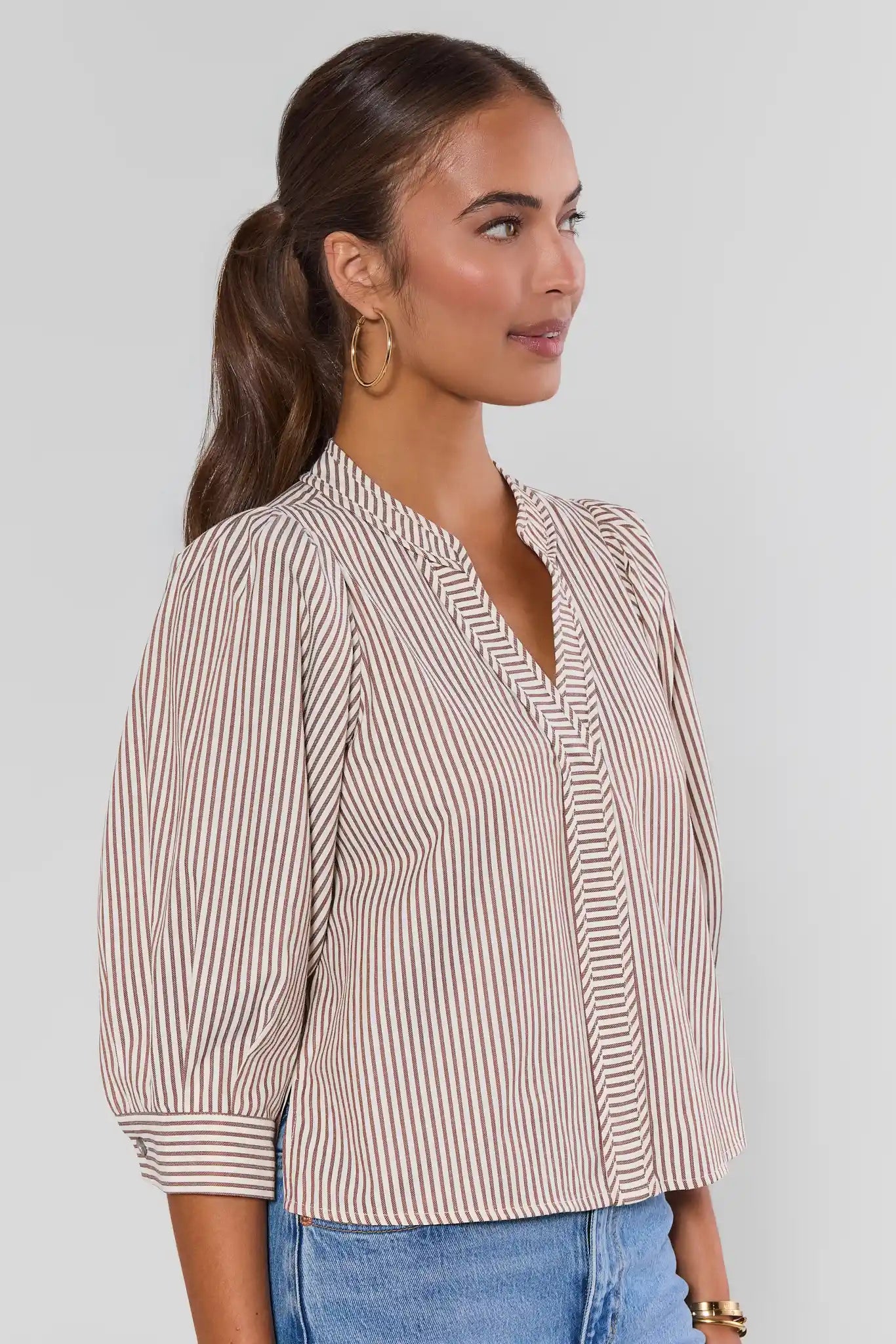 A woman wearing a beige and white vertically striped blouse with puff sleeves and a split V-neckline, paired with blue jeans against a light gray background.